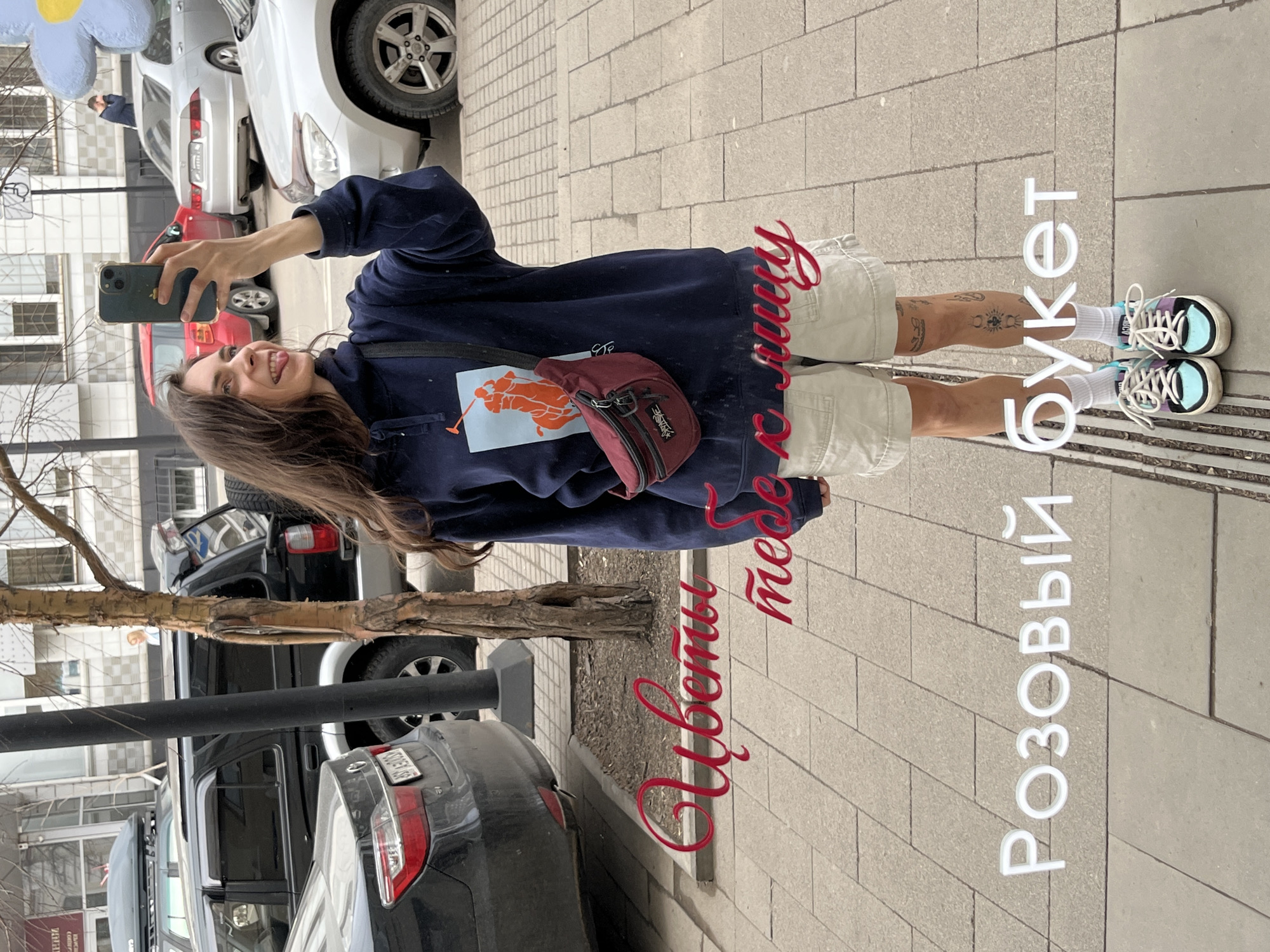 A young woman stands on a sidewalk in the city of Krasnoyarsk, Russia, taking a selfie with her phone. She wears a dark blue hoodie, khaki shorts, and sneakers with blue and purple accents. A red crossbody bag is slung across her torso. Tattoos are visible on her legs. Cars are parked along the curb behind her. A small tree with bare branches stands between her and the vehicles. In the background, apartment buildings are visible. Text, in both white and red, has been added to the image. The red text reads "Цветы тебе в лицу", and the white text reads "РОЗОВЫЙ БУКЕТ".