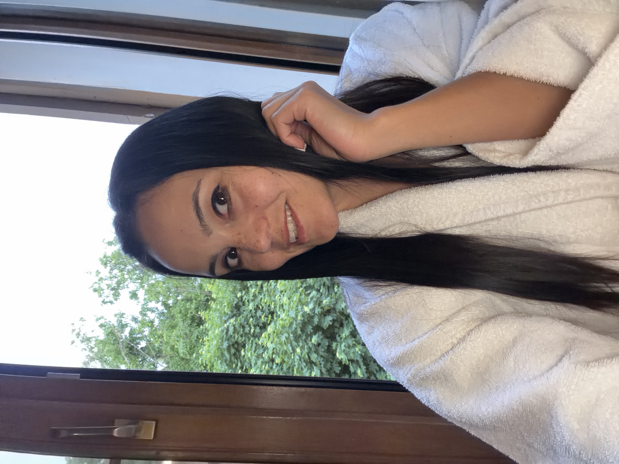 The image captures a young adult female positioned indoors, specifically in the city of Svetlogorsk, Russia. She is situated slightly to the right of the frame's center, facing partially left while smiling directly towards the camera. The woman possesses long, straight, dark brown hair, parted centrally, which cascades over her shoulders. Her complexion is medium, exhibiting visible freckles across her nose and cheeks. Her dark brown eyes are open, with her pupils directed slightly upward and towards the viewer. Her mouth is open in a broad smile, revealing her upper white teeth. She is attired in a white, thick terrycloth bathrobe. Her left hand (viewer's right) is raised, fingers slightly splayed, and positioned near her left temple and hair, suggesting a gesture of touching or running fingers through it. Her fingernails are short and appear to have a light-colored or clear polish.
The background primarily features a window offering a view of the outdoors. On the left side of the image, a dark brown wooden window frame with a brass or brushed metal handle is visible. On the right, sections of lighter-toned wooden window frames are partially seen. Beyond the window, dense green foliage, consistent with trees and bushes, fills the midground, indicating a natural or landscaped exterior. Above the greenery, a bright, overcast or hazy white sky is visible. The scene is illuminated by natural daylight entering through the window.