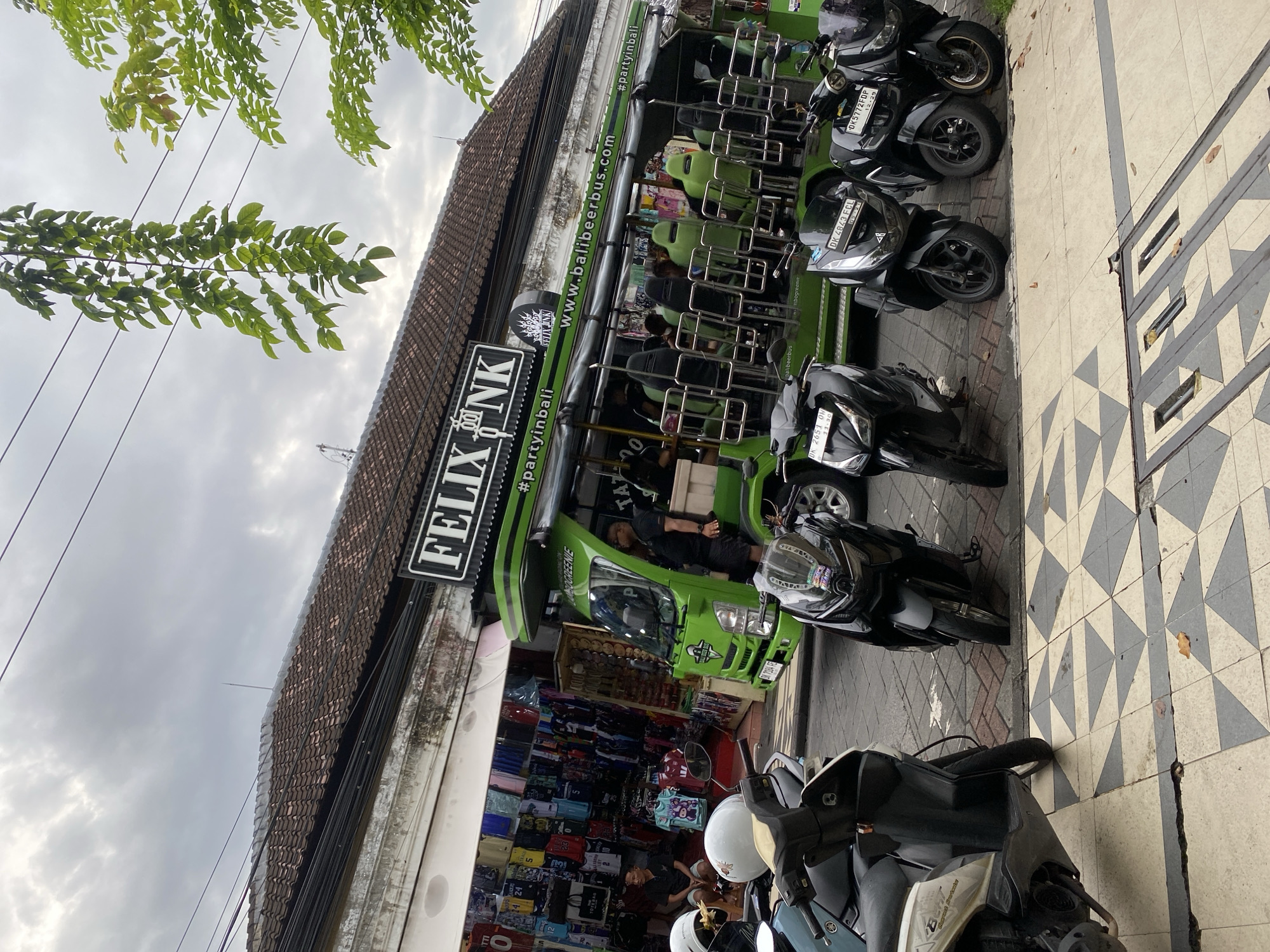 This image captures a lively street scene, likely in Kuta, Indonesia, under an overcast sky during daytime. The perspective is from a slightly elevated viewpoint, looking along a street lined with commercial buildings.
A prominent feature is a bright green, open-sided "party bus" named "FELIXNK" parked along the curb on the right. This bus, labeled with "www.balibeerbus.com" and "#partyinbali", features multiple rows of light green bucket seats with metal railings, and a roof. One person is visible seated inside the bus, and another appears to be in the driver's seat. Adjacent to the bus and extending onto the sidewalk, several black motorbikes and scooters are parked. Two individuals, partially visible on motorbikes in the foreground, are wearing helmets. These motorbikes display Indonesian license plates, such as "DK 2651 ON", "DK 4843 FCL", and "DK 5772 FDP".
On the left side of the frame, a building with a traditional brown tiled roof houses a shop, displaying various clothes and merchandise. Green foliage from an overhead tree extends into the upper left corner of the image, while utility lines crisscross the cloudy sky. The sidewalk features a distinctive black and white patterned tile design. The overall scene suggests a busy tourist-oriented area. Other visible text includes "OGREENIE" and "TATOO" on the bus.