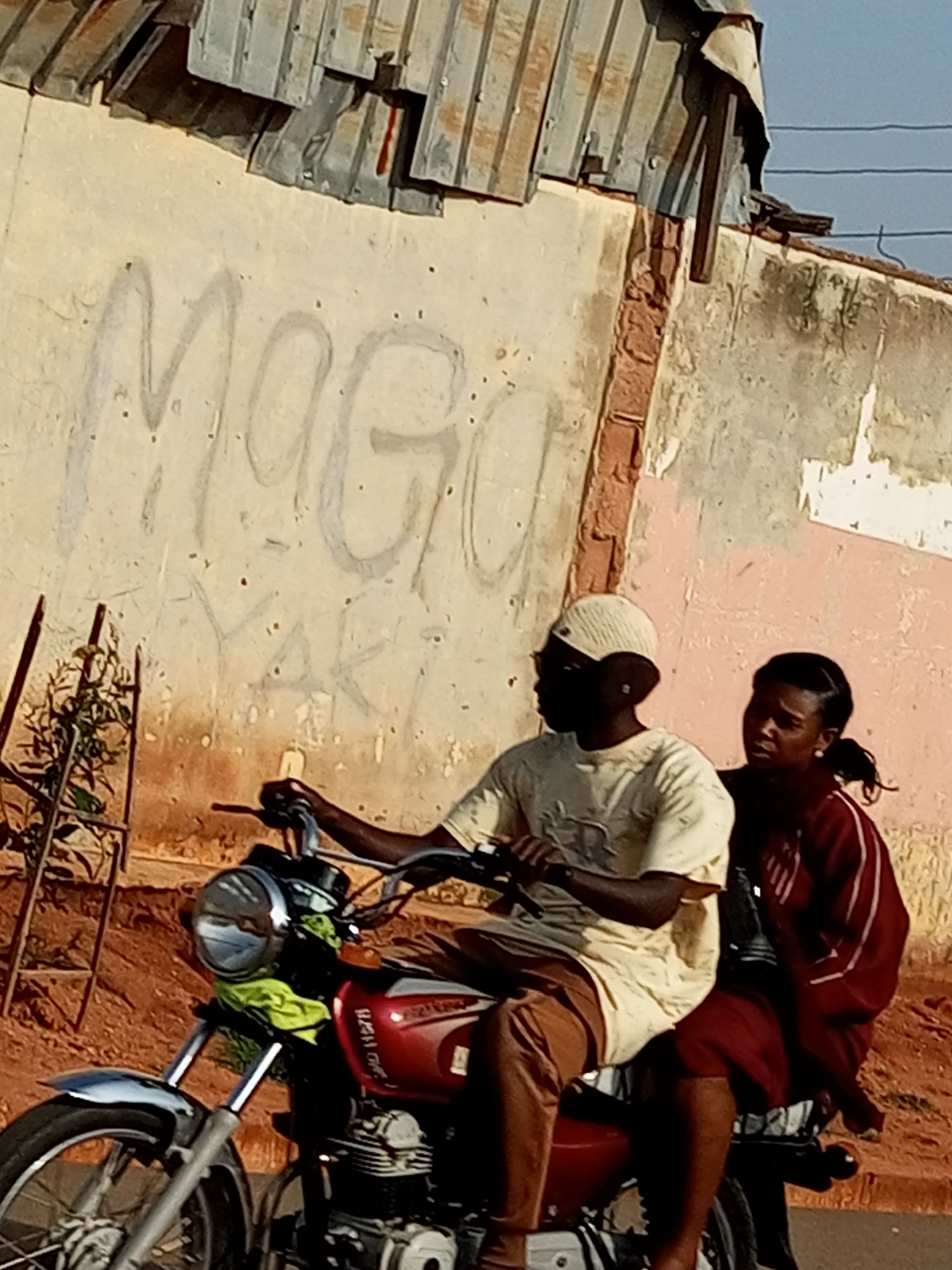 Two individuals, a male driver and a female passenger, are riding a red motorcycle on a dirt road in Tumfure, Nigeria. The male driver, wearing a light-colored shirt and a white cap, is seated in front, operating the motorcycle. The female passenger, dressed in a maroon jacket with white stripes and a maroon skirt, is seated behind him. The motorcycle has a chrome headlight and a green cloth draped over its front. The background features a textured wall with graffiti and a corrugated metal roof. A metal structure with plants is visible to the left of the motorcycle. The sky is clear and blue.