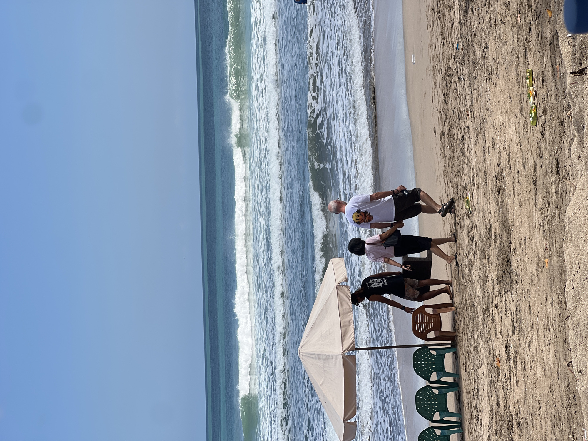 In Kuta, Indonesia, on a sandy beach, several individuals are walking by the ocean under a bright, clear sky. A beige beach umbrella stands on the sand, shading a few plastic chairs, two of which are green and one is brown. Three people are walking away from the camera, towards the ocean. The person in front, on the left, is a man wearing a black t-shirt with graphics and shorts. To his right is a woman in a pink shirt and dark skirt, holding a black purse. Behind them, a man in a white t-shirt with graphics and dark shorts walks with his arm extended. The ocean displays rhythmic waves crashing on the shore, with turquoise water transitioning to a deeper blue horizon. Scattered on the sand in the foreground are some discarded items, including small green offerings with flowers.