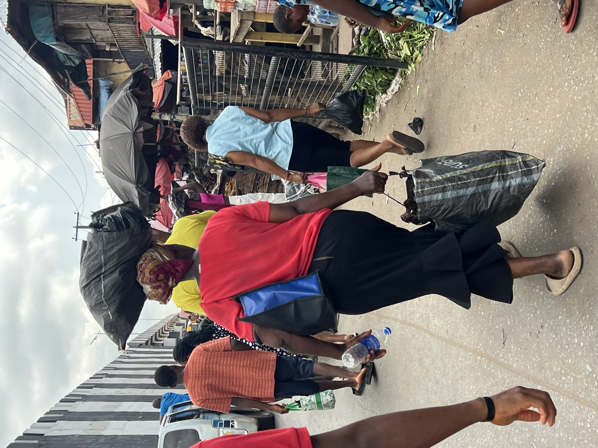 This image depicts a bustling, outdoor market or street scene in Nkpor, Nigeria, under an overcast daytime sky. Numerous people, including adults and children, are seen walking along a paved pathway or bridge, suggesting a busy thoroughfare.
Many individuals are carrying large, heavy-looking black bags, likely containing market purchases, goods, or refuse. In the foreground, a person in a red shirt and black skirt walks, carrying a blue and black shoulder bag. Nearby, a person holds a plastic bottle of "Aquafina" water. Further along, another person in a light blue shirt and black shorts also carries a large black bag, on which the word "SUPPLY" is partially visible.
The background reveals a dense market environment with informal stalls and structures. These appear to be constructed from wood, corrugated metal, and various tarps or fabrics, giving a makeshift impression. A large, open black umbrella provides shade over what could be a vendor's stand. Green leafy produce is visible near a metal fence, suggesting a fresh produce section.
A distinctive black and white striped barrier or wall runs along the left side of the pathway. The overall atmosphere is one of active daily commerce and movement within a densely populated, vibrant market area.