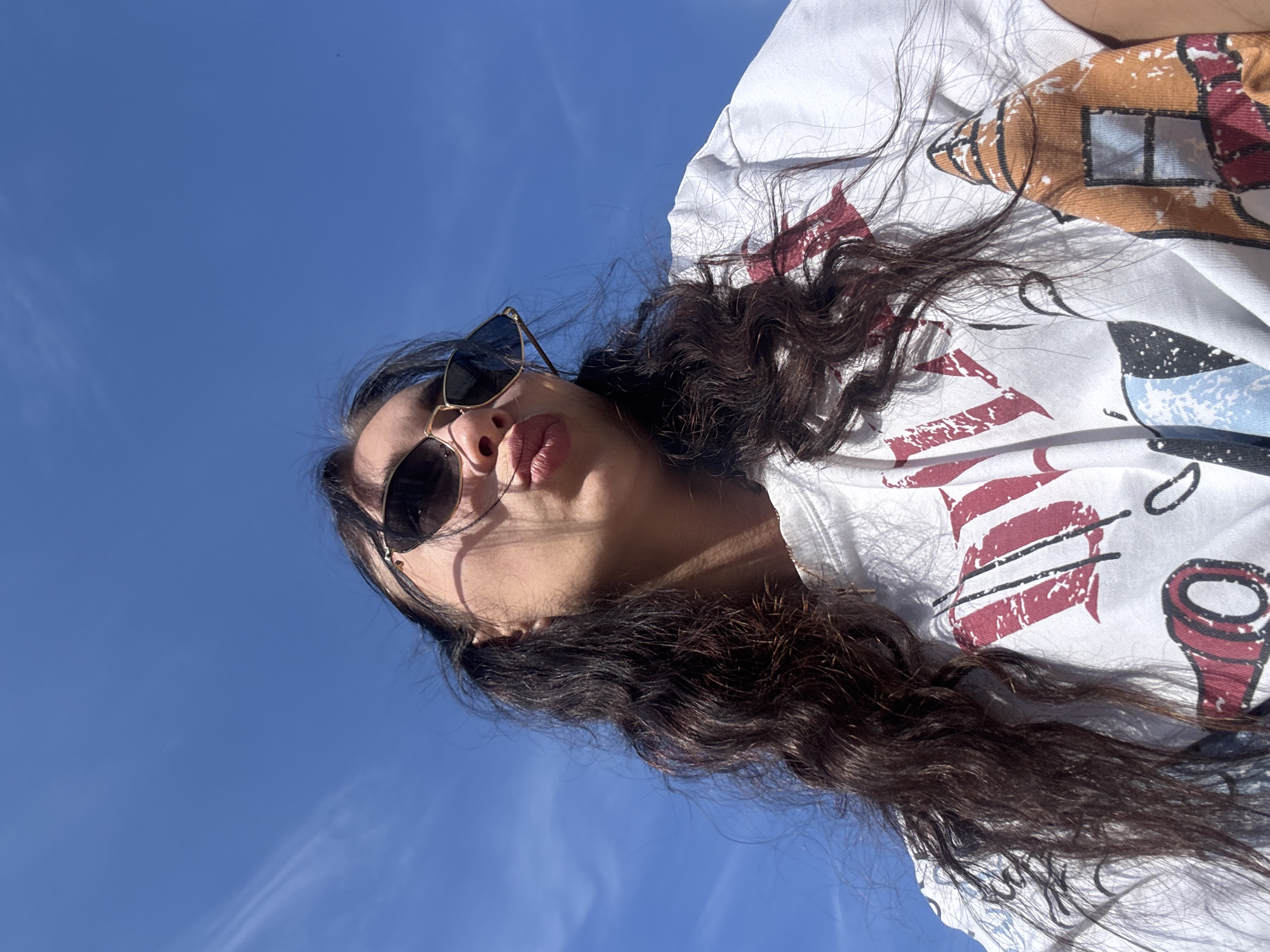 The image captures a low-angle upward perspective of an individual, likely female, against a vibrant, clear blue sky with minimal wispy clouds. The person is facing upwards and slightly to the right, making a playful puckered or "kissing" expression with full, pinkish lips. She wears dark-lensed, gold-framed sunglasses with subtle detailing at the temples, casting a shadow across her upper cheek. Her long, dark brown, wavy hair appears somewhat wind-swept, framing her face and extending down her shoulders. She is wearing a white short-sleeved t-shirt adorned with distressed-style graphic prints. Visible graphics include large red lettering on the viewer's right side of the shirt and a distinct cartoonish brown and orange building-like structure with a blue window on the viewer's left side. Strong, direct sunlight illuminates the scene, creating defined shadows on the person's face and shirt. The background consists entirely of the blue sky. The provided image lacks any discernible visual cues or landmarks to identify the specific location as Chaikovsky, Russia, or any other particular city.