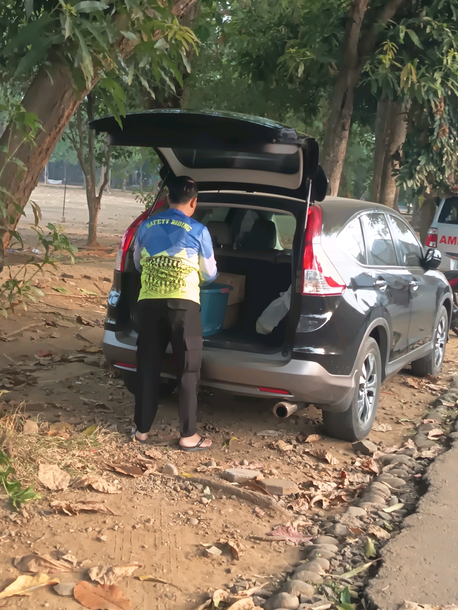 A man wearing a blue and yellow long-sleeved shirt with "SAFETY RIDING" printed on it and black cargo pants stands next to the open trunk of a black SUV. The SUV is parked on a dirt path next to a paved road, with trees and foliage surrounding the area. Inside the trunk, there is a blue bucket and some white plastic bags. The man appears to be putting something into the trunk. The scene is outdoors and seems to be in a park or wooded area.
