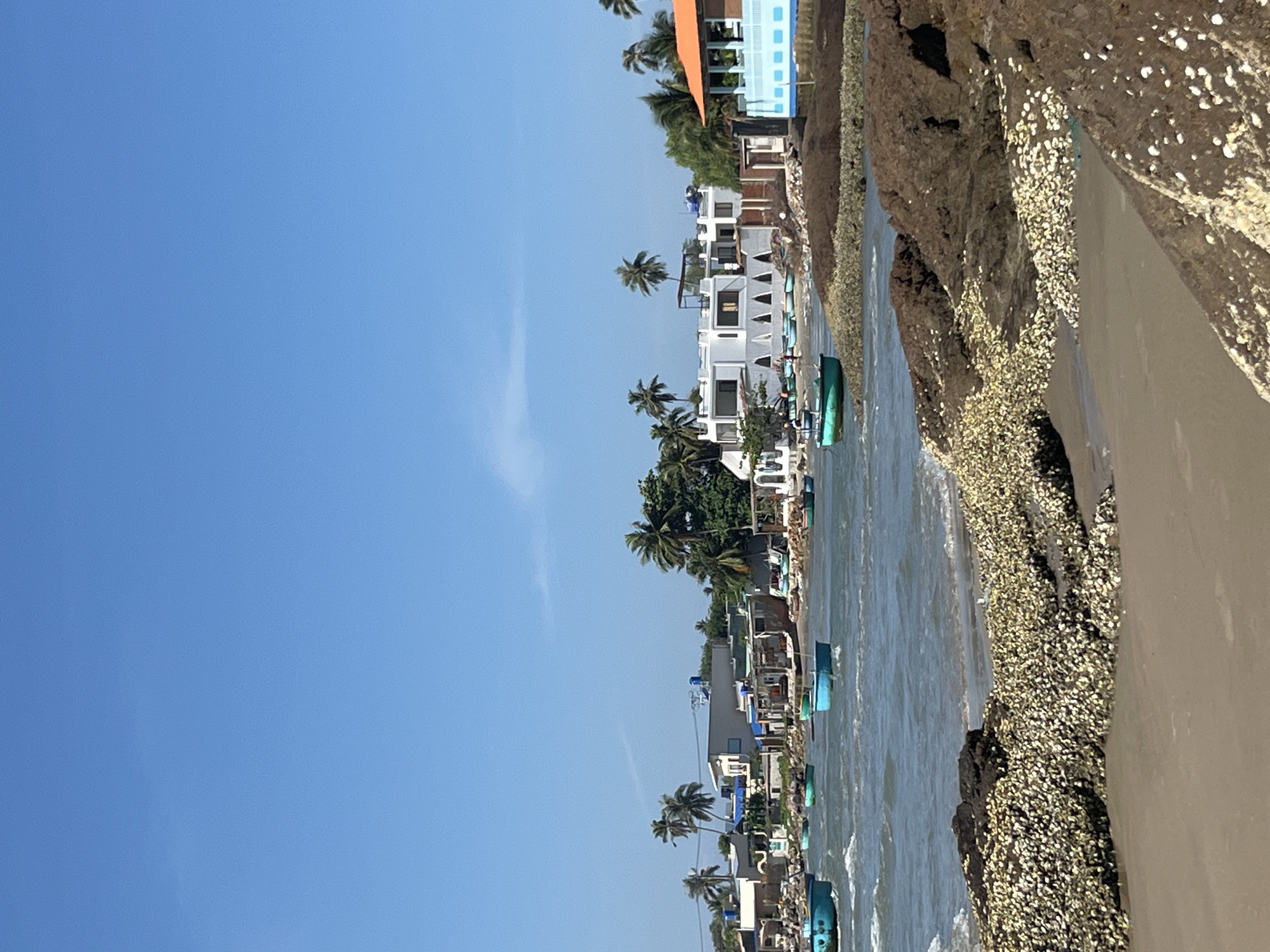 This is a medium-shot, eye-level, vertical, outdoor, daytime, natural light photograph of a coastline in Mũi Né, Vietnam. The sky is clear and blue. In the foreground, the ocean laps onto a sandy beach dotted with large, dark rocks covered in seashells. In the midground, the water is populated by several small, round, turquoise-colored fishing boats. Beyond the water, a shoreline is visible with low-lying buildings, some appearing to be homes or businesses, interspersed with palm trees and vegetation. One building is white with a flat roof and many windows. Another structure to the right has an orange roof and blue and white siding. There are no people clearly visible in the image.