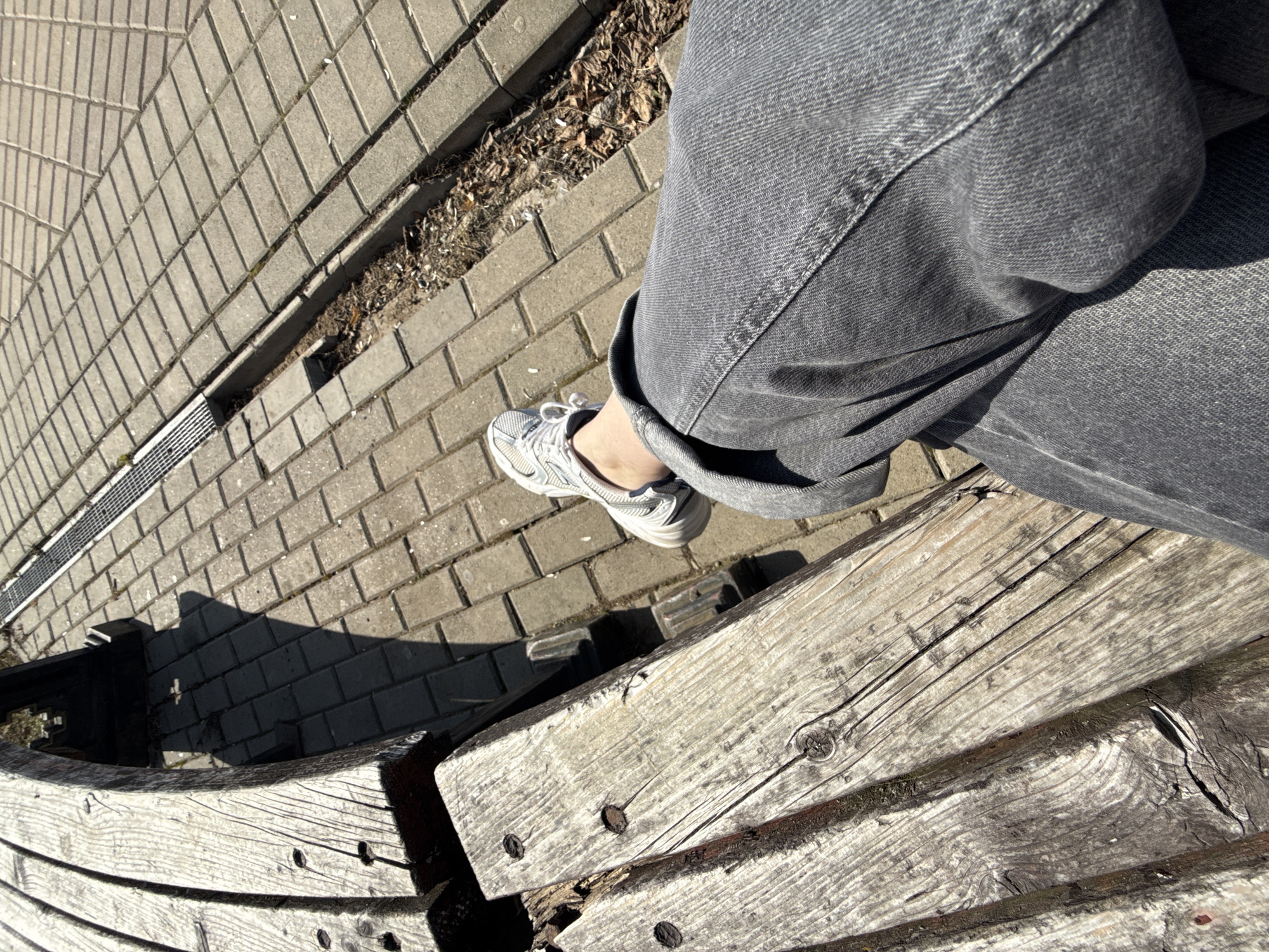 The image depicts a person sitting on a weathered wooden bench with gray denim jeans and a white sneaker visible. The bench is situated on a brick-paved area, with a drainage ditch and dry foliage along the side. The location could be in Izhevsk, Russia, but that cannot be verified from the image alone.