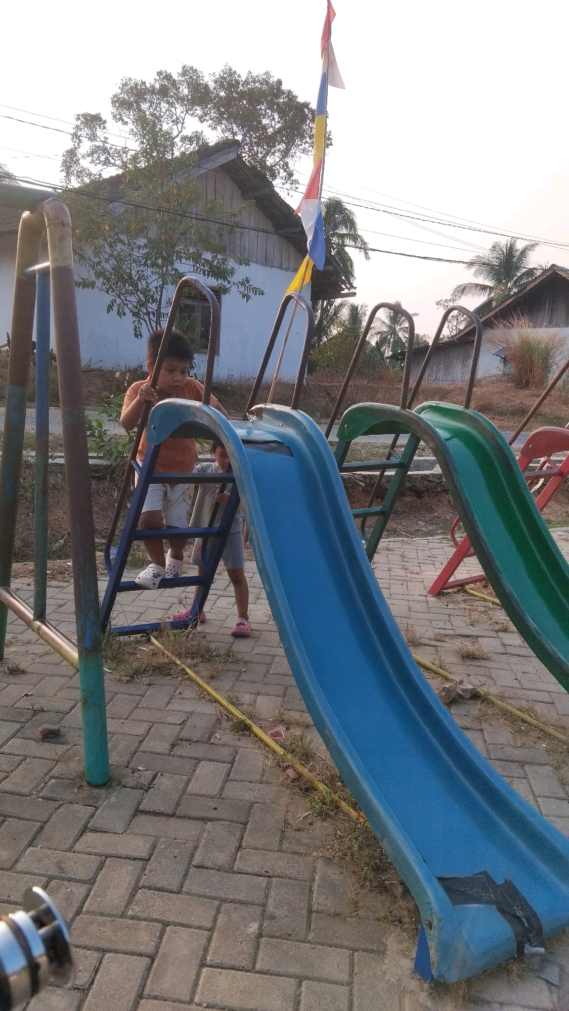 In Manunggal Jaya, Indonesia, a public playground features a blue plastic slide with metal supports, adjacent to a green slide and a red slide support. Two children are present on the playground. A young boy in an orange shirt and white shorts, wearing white Crocs, is climbing the ladder of the blue slide. Behind him, another child, partially obscured, is also near the slide. The playground is situated on a paved surface made of interlocking bricks, with patches of dry grass and dirt surrounding the equipment. In the background, several modest houses with pitched roofs are visible, some appearing to be under construction or in a state of disrepair. A flag, displaying red, white, blue, and yellow sections, flies from a pole near the houses. Power lines are strung across the sky, and palm trees are visible amongst the houses, indicating a tropical or subtropical climate. The overall scene depicts a simple, outdoor play area in a semi-rural or residential setting.