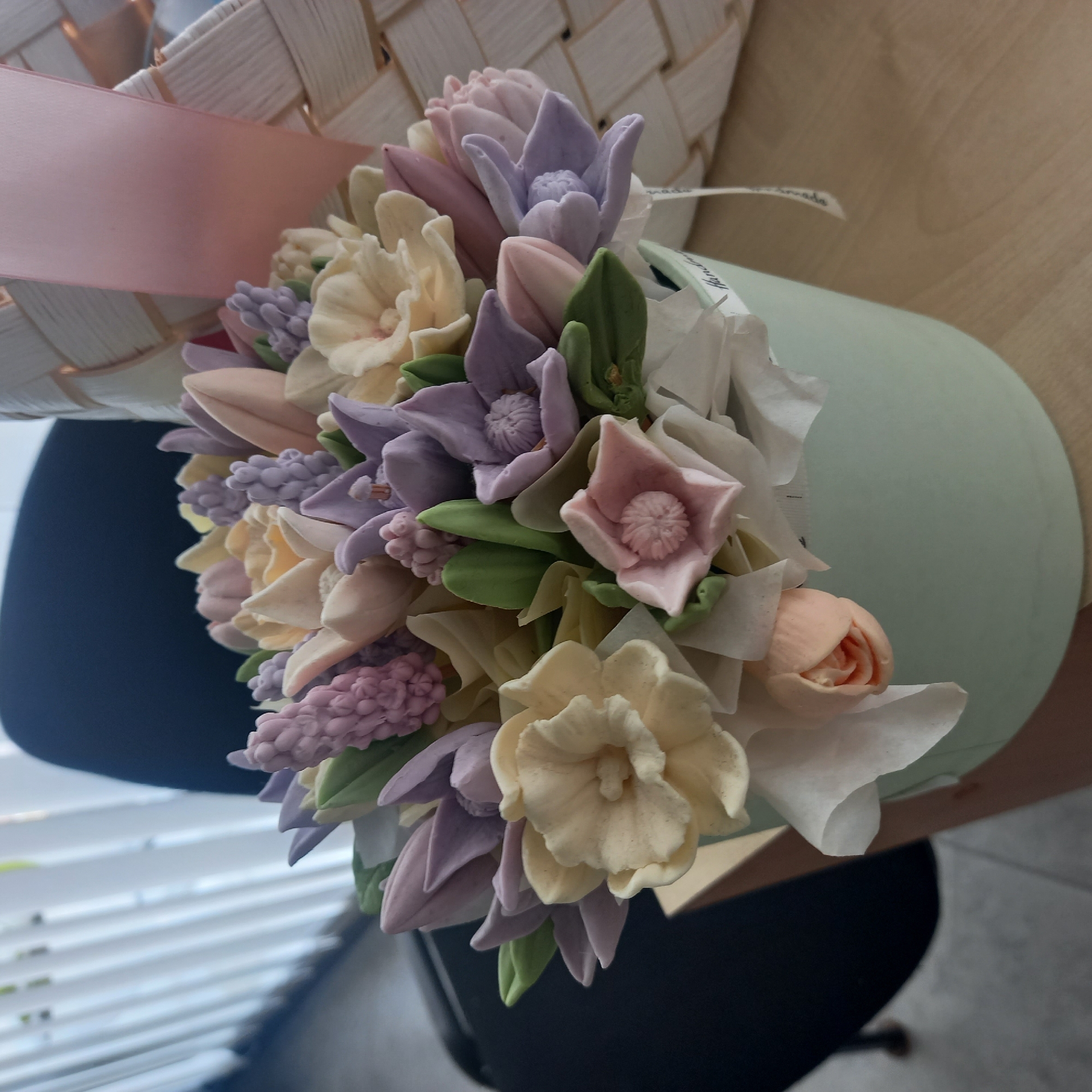 A close-up, high-angle shot captures a meticulously crafted bouquet of artificial flowers arranged in a pale green cylindrical container. The flowers are made of a delicate material, possibly fondant or frosting, and come in a pastel palette of soft yellows, lavenders, pale pinks, and light peaches. Among the blossoms are textured clusters resembling hyacinths, and petal designs that mimic tulips and daffodils. Green fondant leaves are interspersed throughout the arrangement, adding a touch of natural contrast.
The bouquet is partially cradled within a woven basket, its texture and color suggesting it might be made of straw or a similar natural material. A pale pink ribbon is draped across the basket, adding a soft decorative element. A small white ribbon with faint text, likely a brand or "handmade" label, is visible attached to the bouquet's wrapping.
The setting appears to be indoors, with the bouquet resting on a wooden surface, possibly a table. In the background, a dark chair is visible, and to the left, a white slatted structure, reminiscent of blinds, suggests a window. The overall lighting is soft and diffused, indicating it might be daytime, though no specific time can be determined. The composition evokes a sense of gentle beauty and artisanal skill. No people are visible in the image.