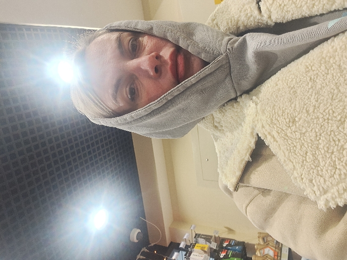 A person in a gray hoodie with the hood up and a sherpa-lined jacket. The individual's face is visible with pale skin and neutral expression. There's a bright glare in the upper left corner, possibly from a light source. The background features a dark, textured wall on the left and beige-colored walls and what appears to be a shelf or counter with items on the right.