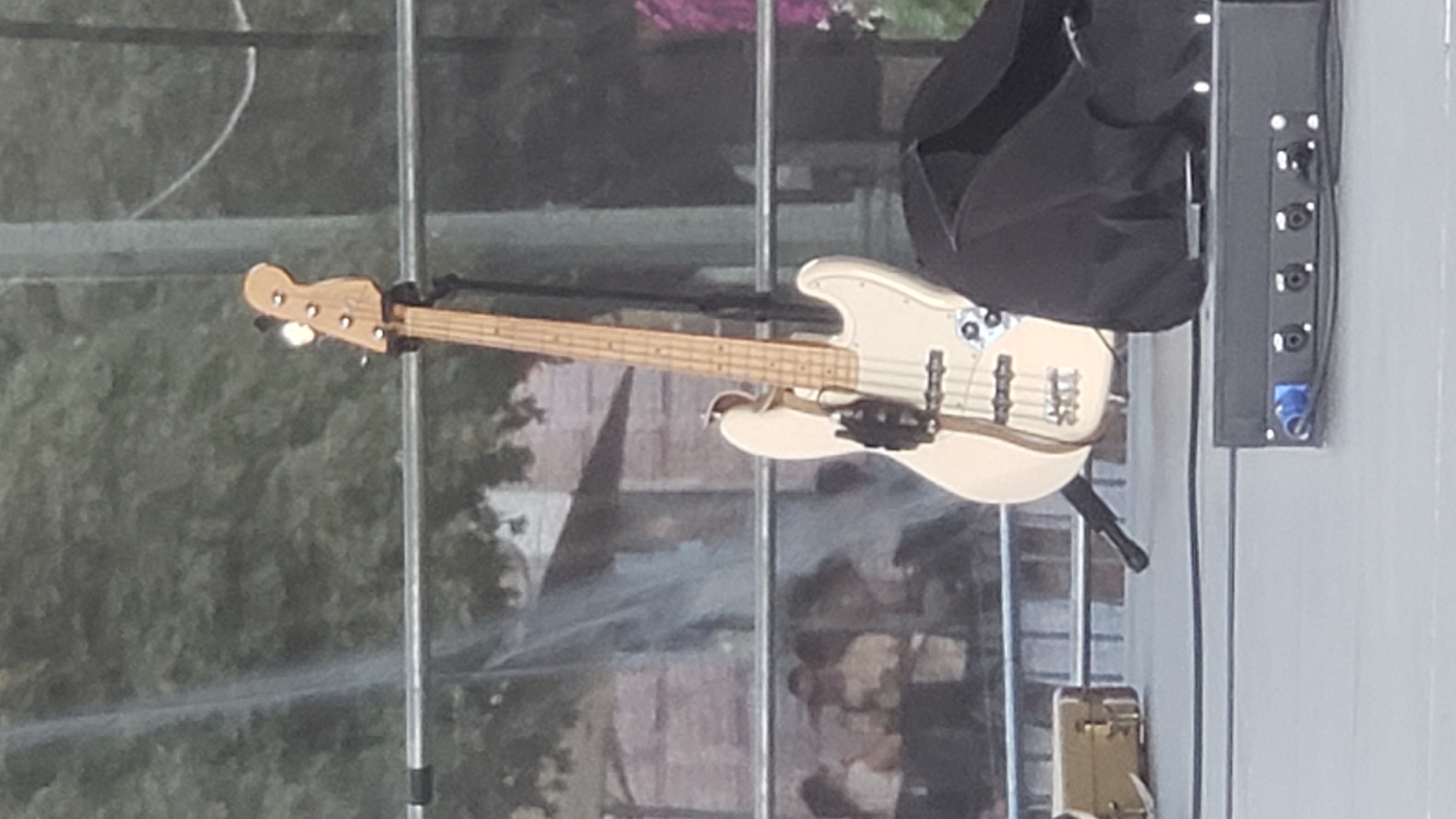 A white electric bass guitar stands upright on a stage in what appears to be an outdoor setting, possibly a concert or festival. The guitar is on a stand, with a black cable draped over it. To the right of the guitar is a black cloth or garment. In the foreground, a black electronic device with several connectors is visible. Behind the stage, a metal railing separates the performance area from a blurred background of trees and buildings, suggesting a public space in a city. There are glimpses of people in the background, some appearing to be standing or walking. The overall lighting suggests it is daytime, but the specific location in Prešov, Slovakia cannot be determined from the image alone.