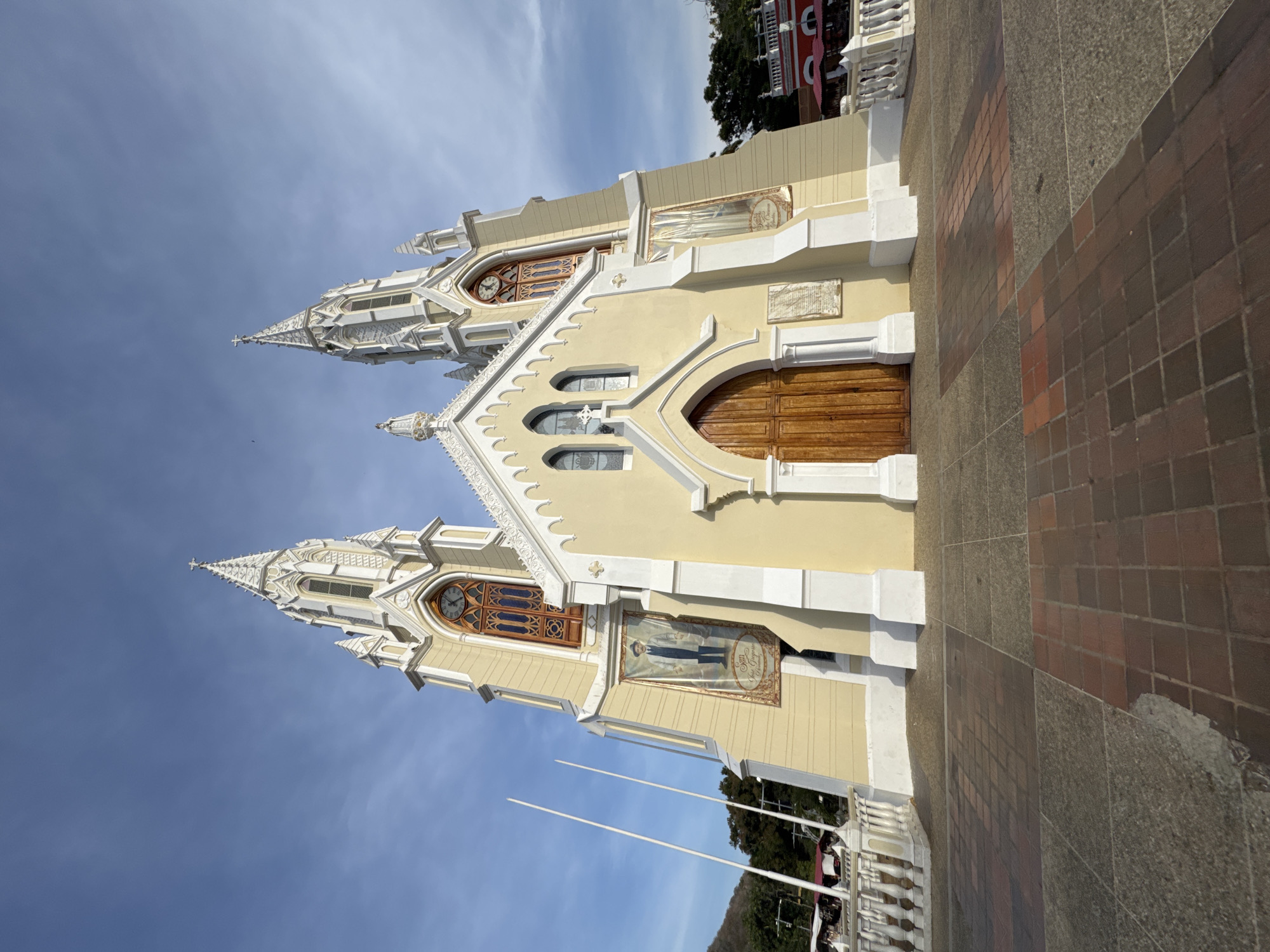 The image showcases a majestic, light yellow church with intricate white architectural detailing, photographed from a low angle under a bright blue sky with wispy clouds. The church features two prominent, ornate spires topped with crosses, each housing slender windows and decorative elements. The main facade presents two large, arched wooden entrance doors. Above the doors, several smaller arched windows with grille work are visible, and further up, a large rectangular window with a clock face is set into one of the towers.
A notable detail on the church facade is a large vertical banner or painting depicting a man in a white suit, identified by the visible text "San José Gregorio Hernández," indicating a depiction of the venerated Venezuelan saint.
The foreground consists of a paved area, likely a plaza or sidewalk, constructed from a mix of brown and grey square tiles. Several white poles, possibly flagpoles, stand in front of the church. In the background, green trees and a distinctive red building with white balustrades can be seen, suggesting a vibrant community setting. The bright sunlight indicates it is daytime, enhancing the architectural features and colors of the building. The scene depicts a significant religious landmark in El Valle del Espíritu Santo, Venezuela.