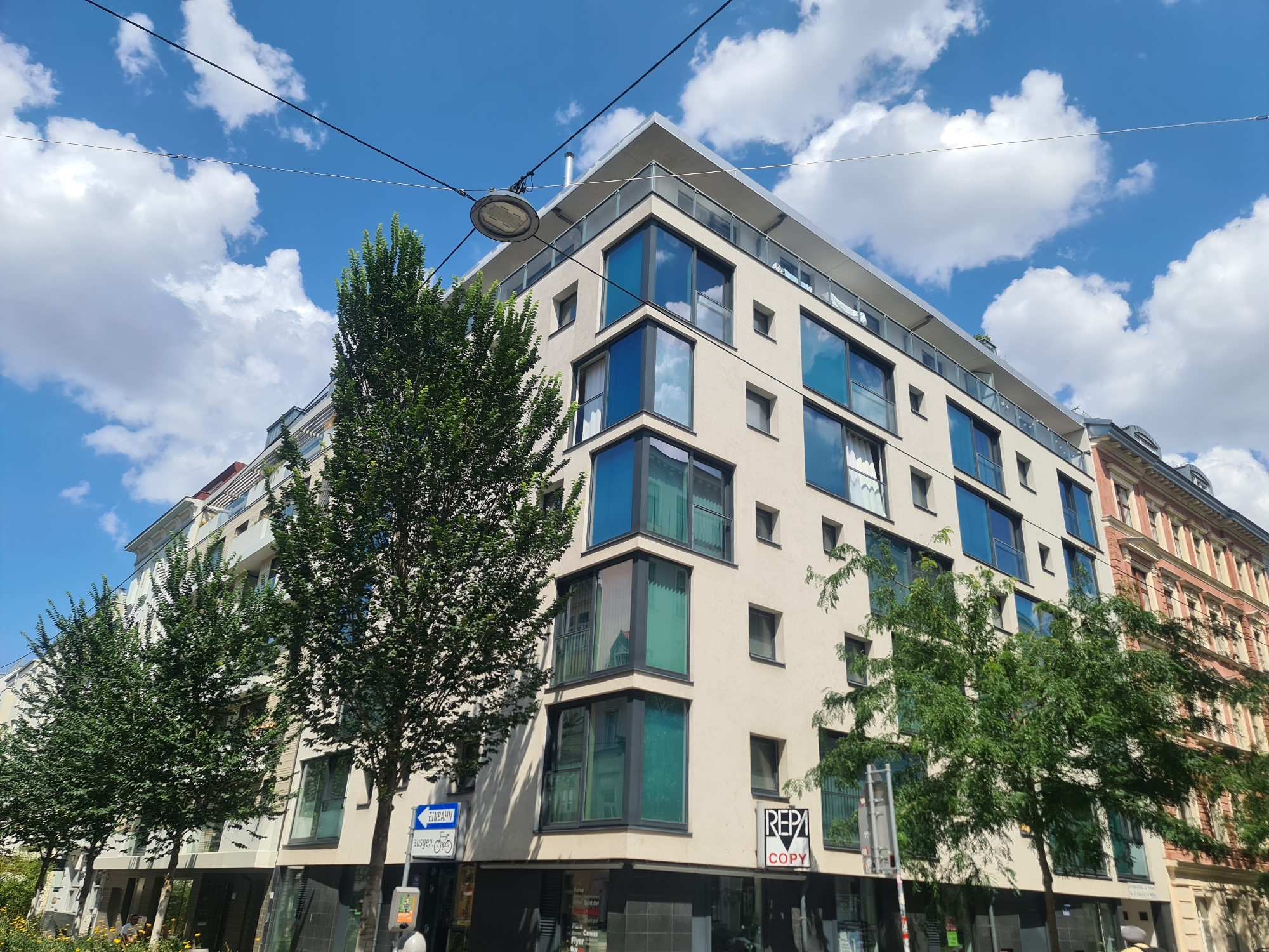 The image shows a modern building with a beige facade and large, blue-tinted windows on the corner of a street in Vienna, Austria. The building has multiple floors, a flat roof with a glass railing, and a contemporary architectural style. Several trees with lush green foliage are planted in front of the building. To the right of the modern building, a section of an older, ornate building with red and cream colored brickwork and arched windows is visible. A street sign indicating "EINBAHN" (one-way street) and a bicycle symbol is posted on the left side of the modern building. The sky is blue with scattered white clouds. The scene appears to be a street view in a city.