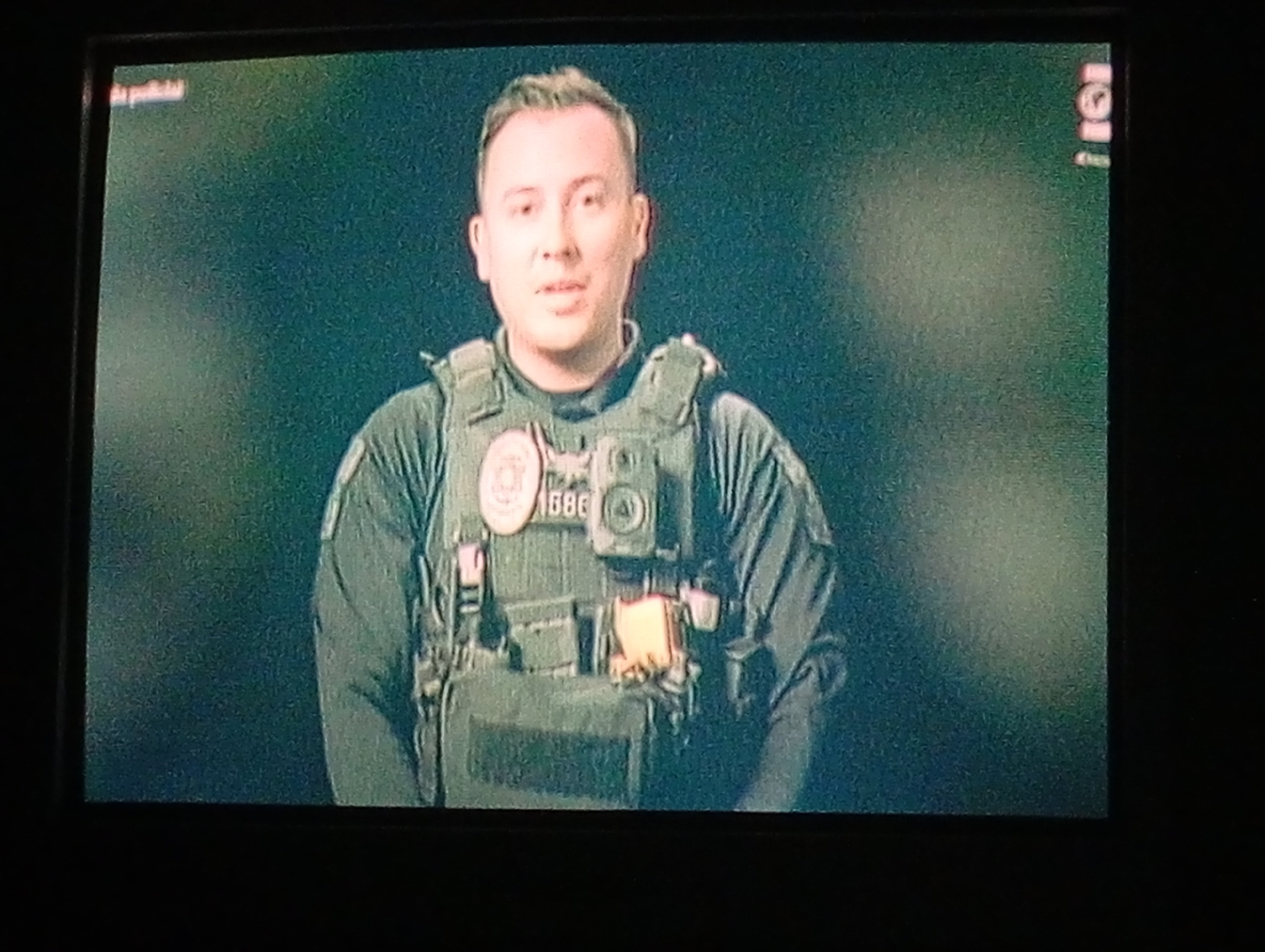 A medium shot shows a man wearing a dark, tactical-style uniform and a ballistic vest. On the vest, a badge and a body camera are visible. The background is a blurry, dark green. The image appears to be captured from a television screen. There is no information to identify Juan Griego, Venezuela.