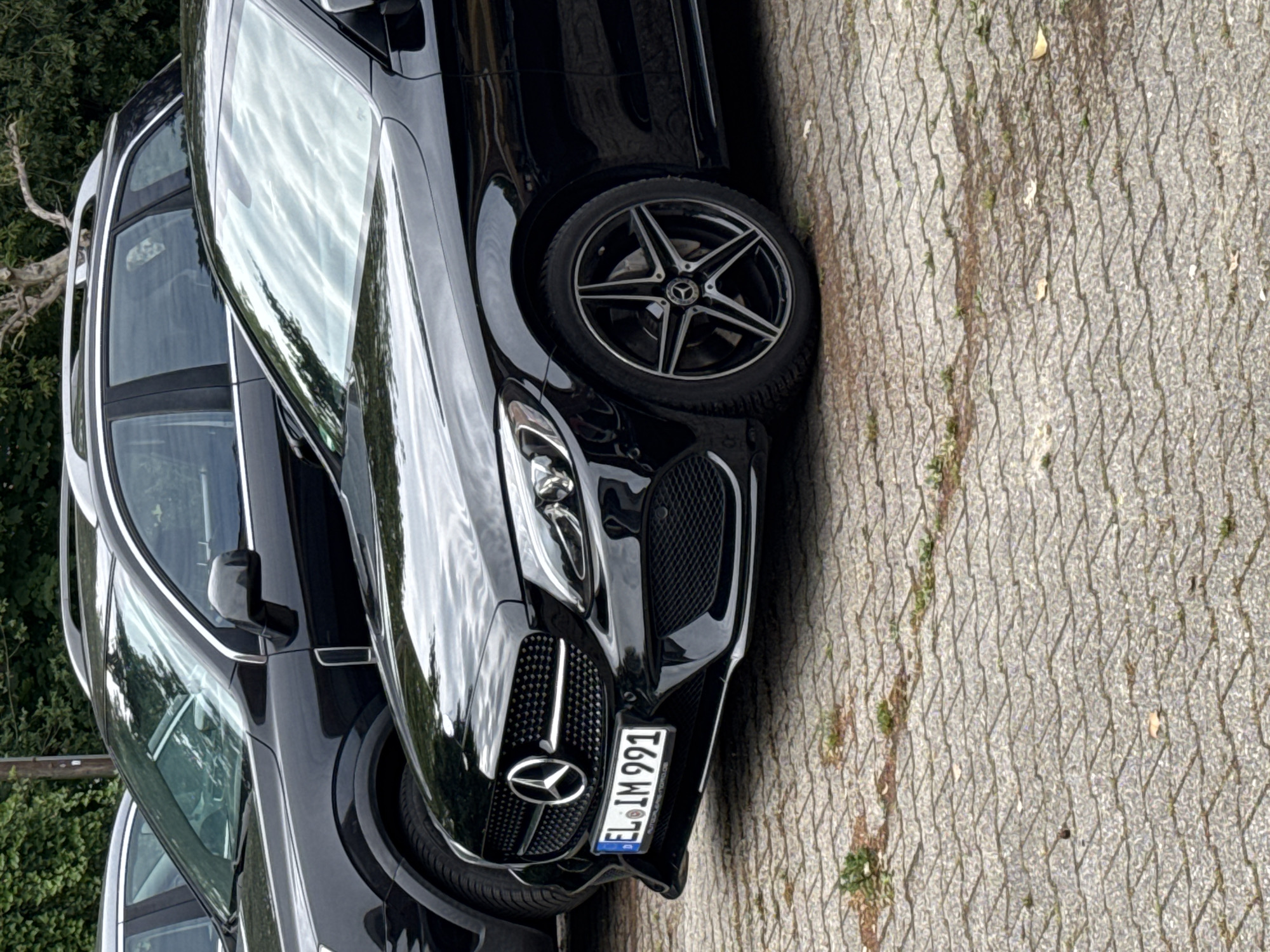 The image captures a black Mercedes-Benz car, likely an SUV or crossover model, parked outdoors on a paved surface. The front right side of the vehicle is prominently featured, displaying its modern headlights, a distinctive grille with the Mercedes-Benz star logo, and a stylish multi-spoke alloy wheel with a center cap also bearing the Mercedes emblem. Reflections of a bright, possibly overcast sky are visible on the car's glossy black paintwork and windows, indicating daytime. A second dark-colored vehicle is partially visible parked behind the main car.
The ground is made of light grey, textured pavers, interspersed with minor dirt, stains, and small patches of green vegetation in the cracks. Green foliage and trees are visible in the background on the left, suggesting a natural or residential backdrop.
A German license plate is affixed to the front of the Mercedes, clearly displaying the characters "EL IM 991" and the "D" country code on the blue Euroband. Small, unreadable text, possibly a dealership name beginning with "AUTOHAUS," is also visible below the main numbers. The "EL" district code on the license plate corresponds to Emsland, which includes Twist, Germany, the provided location context. No people are visible in the scene. The cars appear to be stationary, parked on the surface.