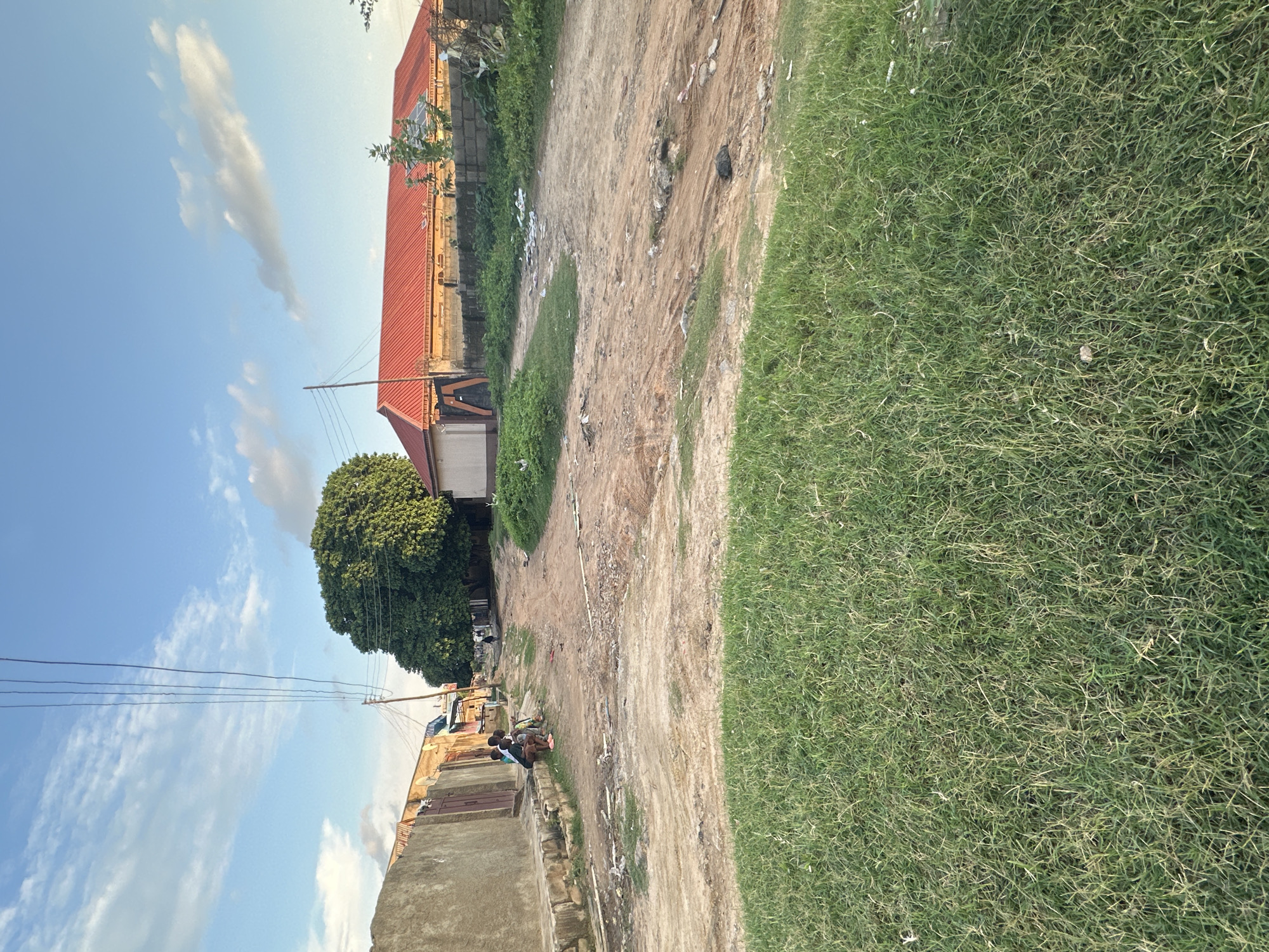 The image depicts a street scene in Narayi, Nigeria, with a dirt road winding through the frame. In the foreground, a patch of green grass is visible. The road is flanked by buildings, including one with a red corrugated metal roof. Several people are present, including a group of individuals sitting on a low wall on the left side of the road and a lone figure walking in the distance. Electrical wires hang overhead, supported by poles. The environment appears somewhat unkempt, with debris scattered along the roadside. The sky is blue with scattered clouds.