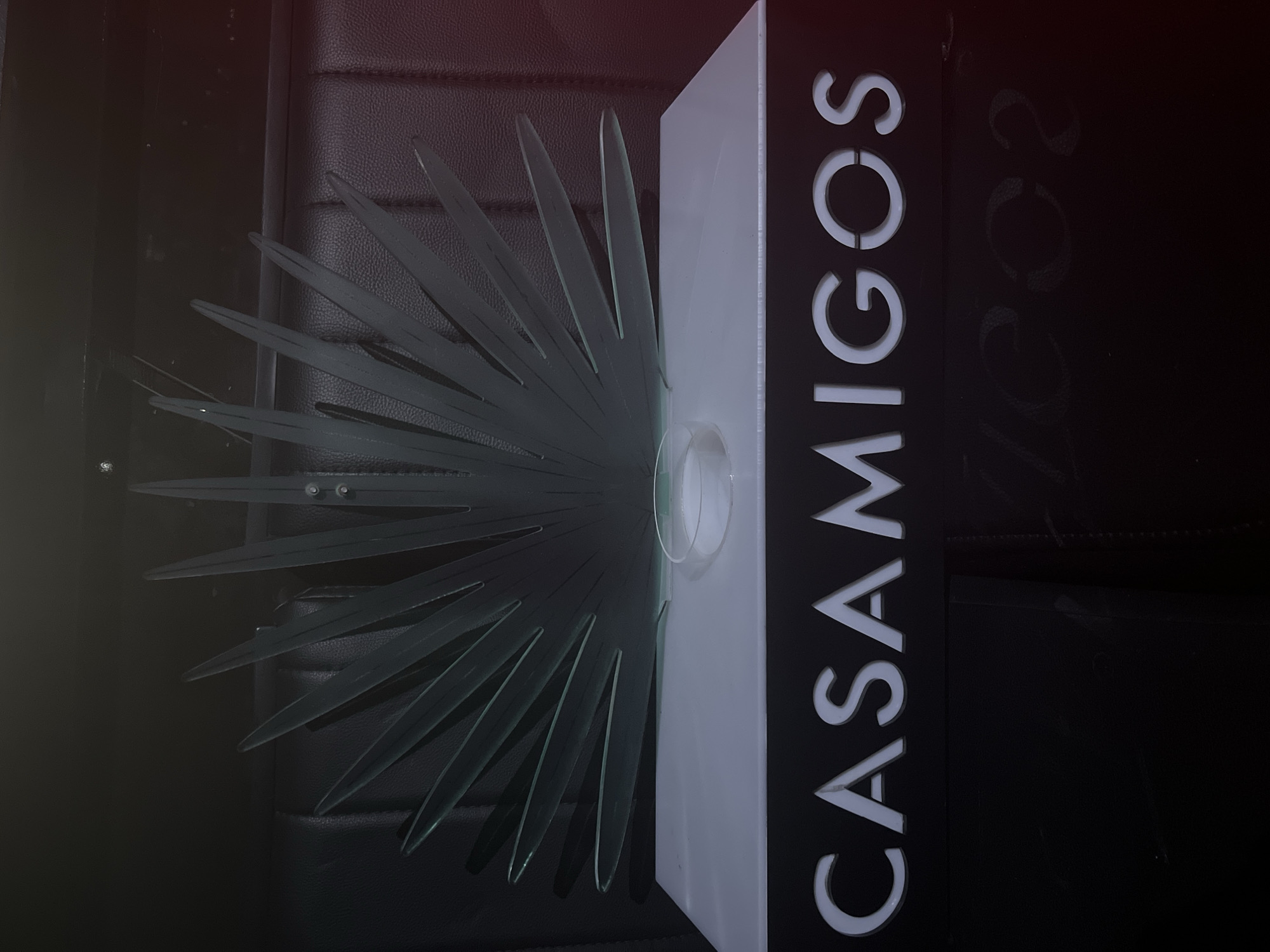 The image shows a decorative item, likely a sign or a centerpiece, placed on a dark, textured surface that appears to be leather. The object itself is comprised of two main parts. On the left, there is a fan-like structure made of clear, possibly glass or acrylic, material. This structure has multiple elongated, pointed segments radiating outwards. In the center of this fan, there is a small, cylindrical clear container.
To the right of the fan is a vertical rectangular plaque. The front of this plaque is black with white lettering that spells out "CASAMIGOS" vertically. The letters are stylized and appear to be cut out or raised. On the black surface to the right of the "CASAMIGOS" lettering, there is faint, chalk-like writing that is difficult to fully decipher but appears to include the numbers "2002".
The lighting in the image is dim, with a reddish tint in the upper left corner, suggesting artificial lighting or a specific time of day. The overall mood is somewhat mysterious due to the lighting and the dark background. There are no people visible in the image. The setting appears to be indoors, possibly in a bar, restaurant, or a private establishment, given the materials and the branded text. The context "Howayi, Nigeria" indicates the geographical location where this image was taken. The "CASAMIGOS" text suggests a connection to the tequila brand of the same name.