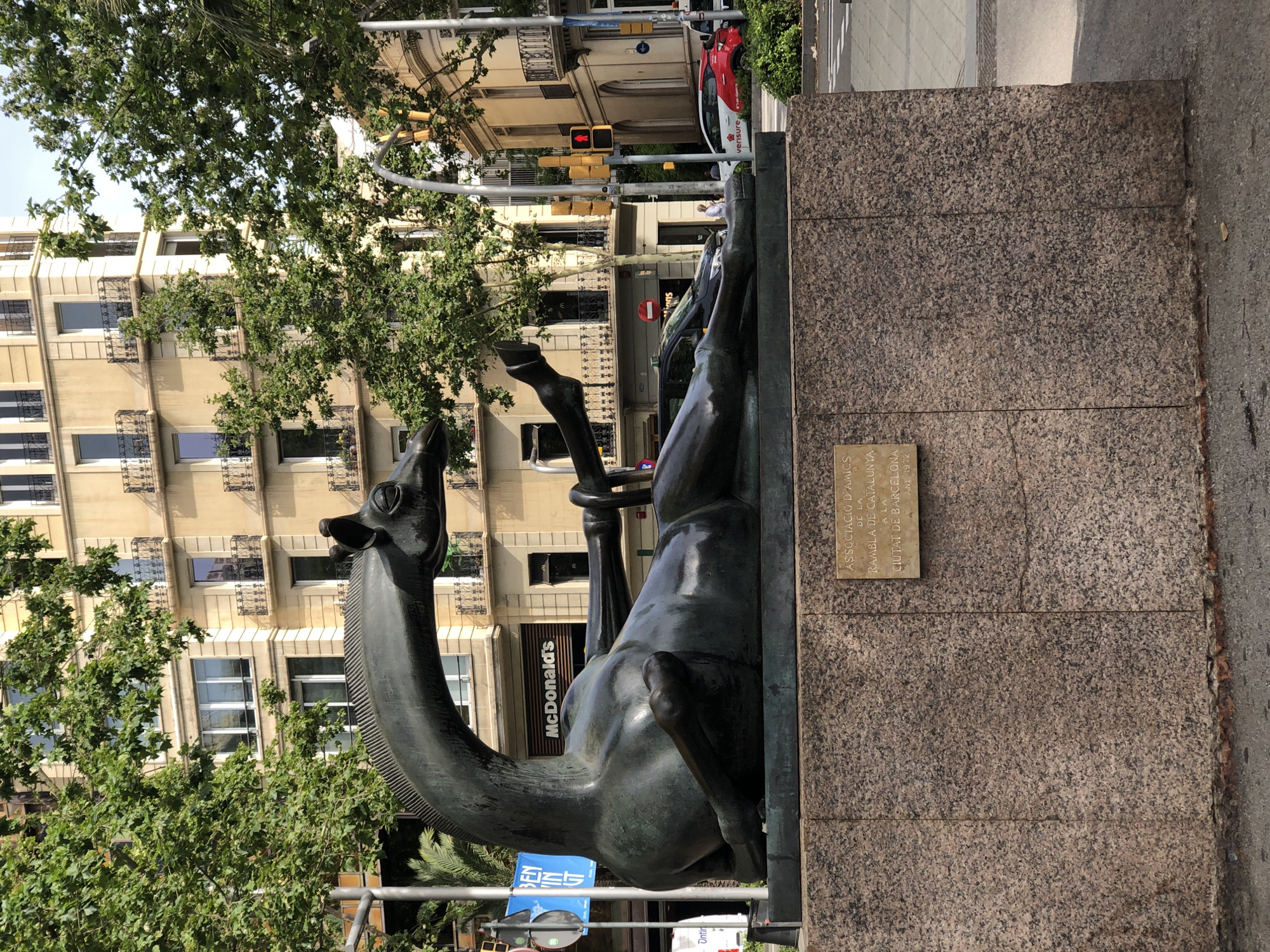 A bronze sculpture of a giraffe is displayed on a granite pedestal in front of a tan building with balconies. The giraffe is lying down, with its head resting on its back and its legs extended. A plaque on the pedestal reads "ASSOCIACIO D'AMICS DE LA RAMBLA DE CATALUNYA A LA CIUTAT DE BARCELONA ANY 1972". In the background, a McDonald's restaurant is visible, along with a traffic light and several cars. Trees and foliage are present in the scene.