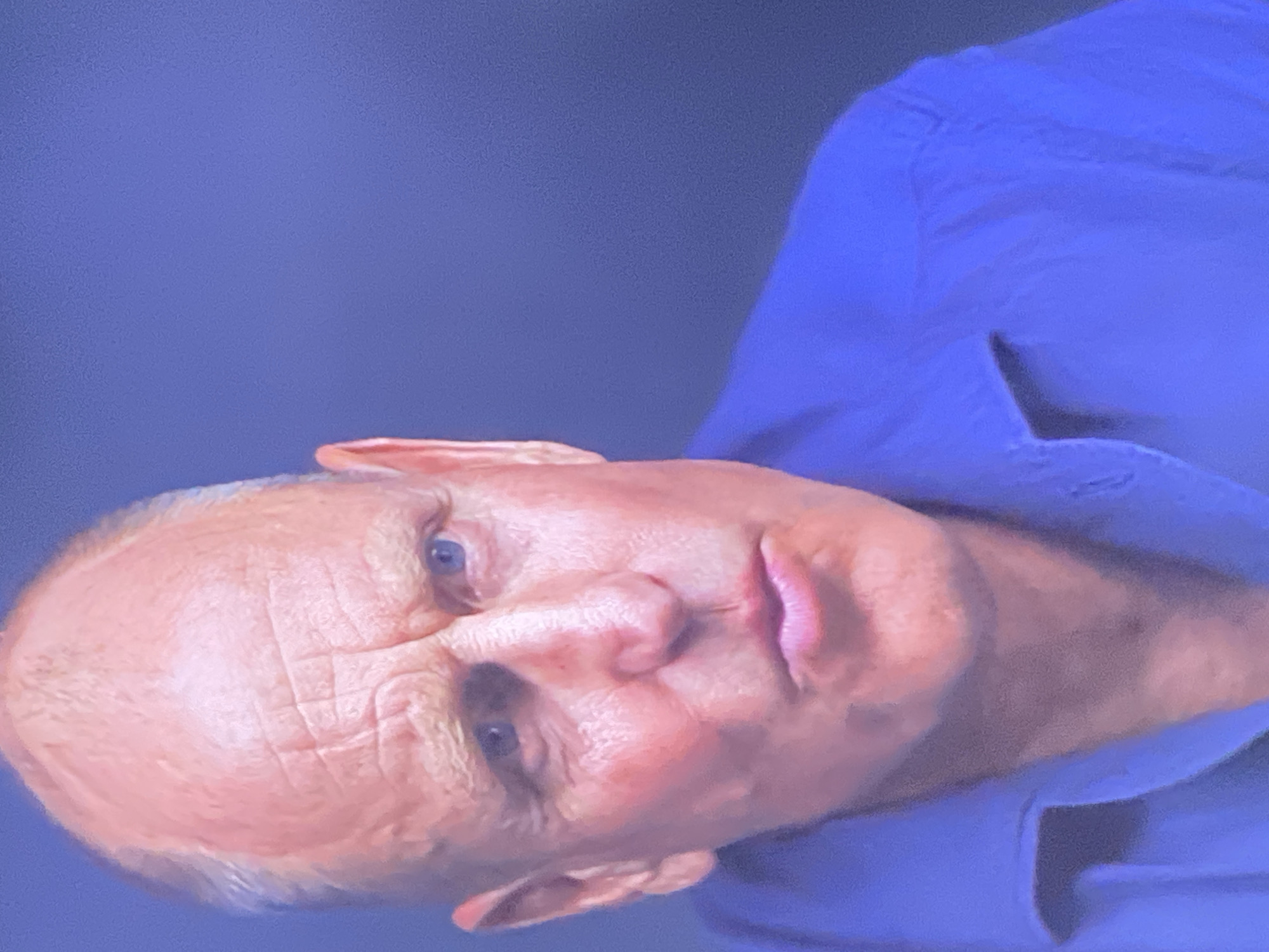 A close-up, head-and-shoulders shot shows a light-skinned man with a shaved head. He is looking slightly to his right and has a serious expression. He is wearing a bright blue collared shirt. The background is a plain, dark blue, out of focus. There is no indication of the city of Zastavna, Ukraine, or any urban environment in the image.