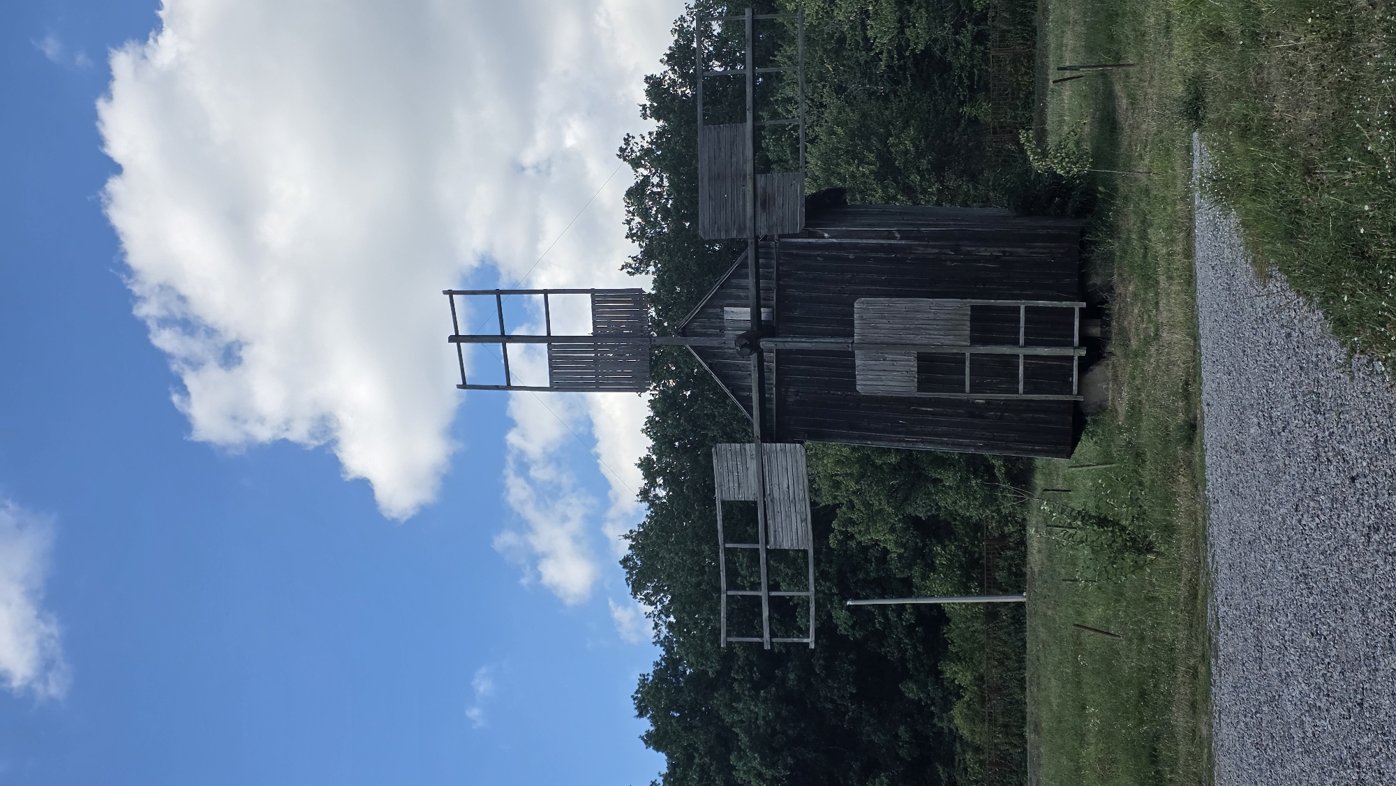 The image depicts a wooden windmill located in a grassy field, surrounded by a dense forest. The windmill is dark in color and appears old. It has two visible wooden blades, one on the left and one on the right, with a simpler structure on top. The sky above is blue with scattered white clouds. The foreground shows a gravel path. The overall setting suggests a rural or historical park environment. The windmill is located in Kyiv, Ukraine.