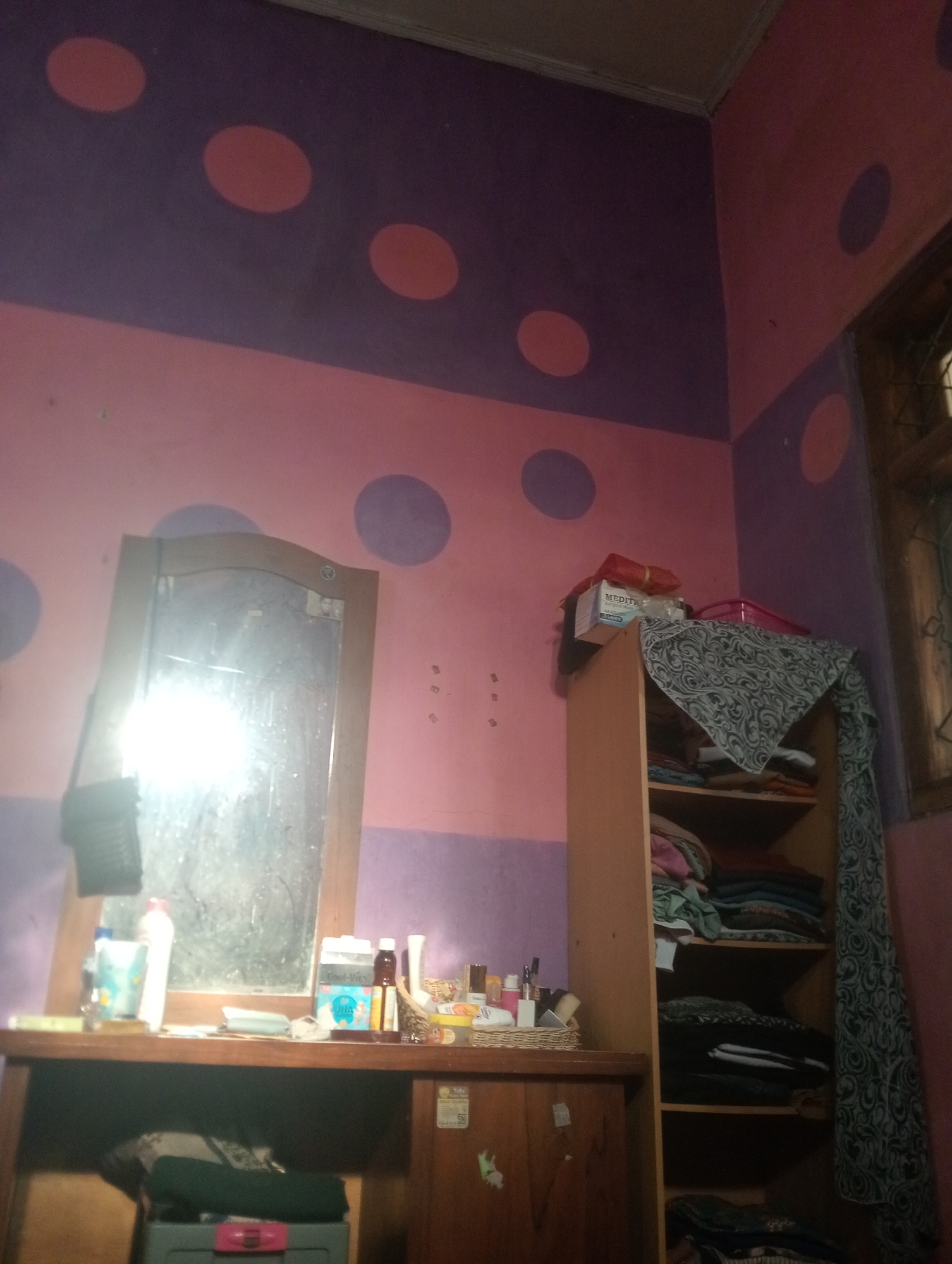 This is a medium shot, indoor photograph of a room with a polka dot wall design in purple and pink. On the left side of the image is a wooden vanity with a mirror, reflecting a bright light source. Various bottles and containers are placed on the vanity. To the right of the vanity is a tall, wooden shelf unit filled with folded clothes. A patterned fabric is draped over the top shelf of the unit. A window is visible on the far right side of the image. The overall impression is that of a bedroom or dressing area. The image was taken in Bloro, Indonesia.