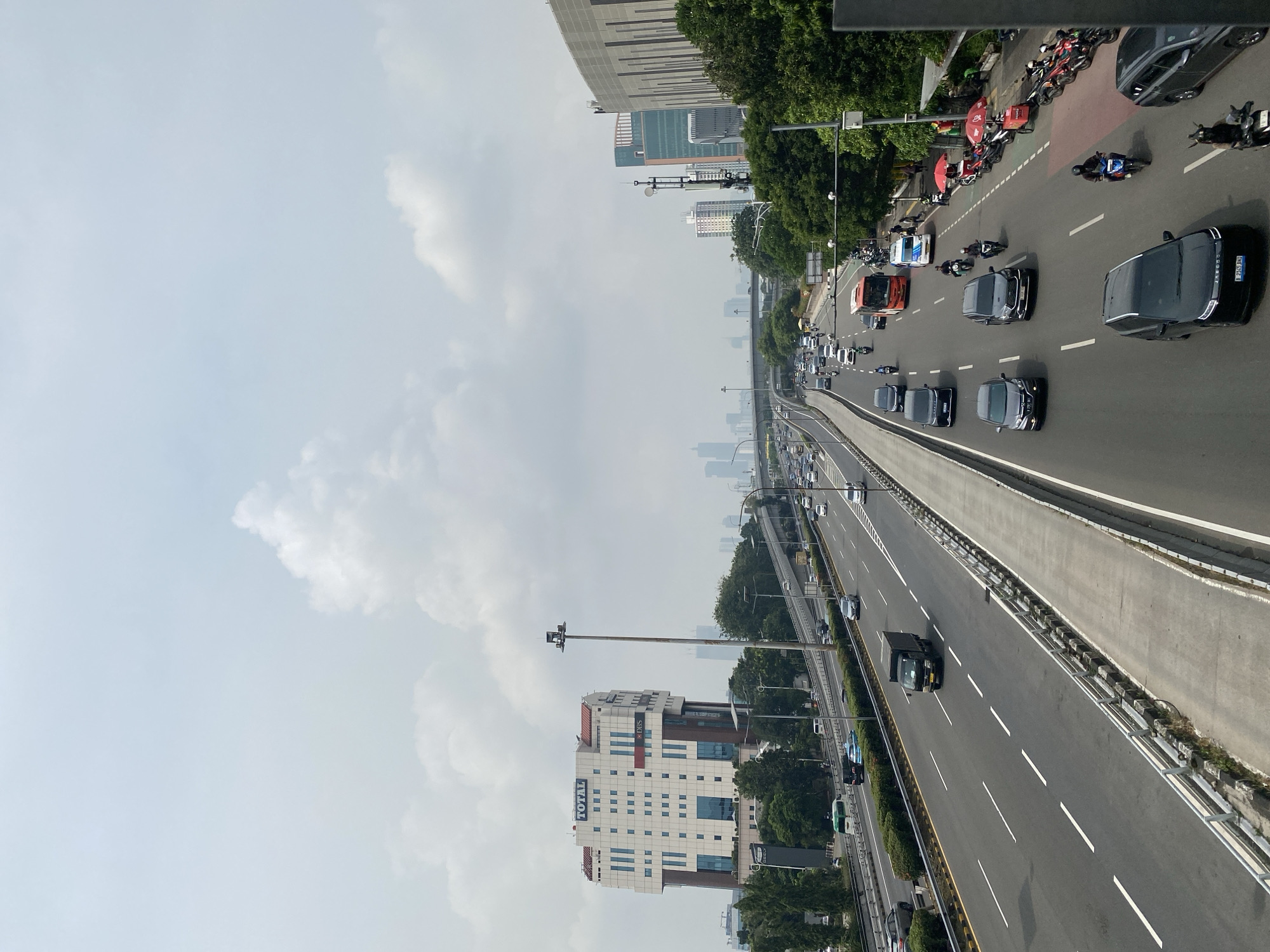 The image captures a wide-angle view of a multi-lane highway in West Jakarta, Indonesia, under a hazy, overcast sky. The highway is busy with traffic, including cars, trucks, buses, and numerous motorcycles. On the left side of the highway, a large, modern office building with the "TOTAL" logo is prominent, featuring a white facade with numerous windows and some blue accents. Adjacent to it, a smaller building with "DBS" branding is visible. Trees and greenery line the sides of the highway, and elevated sections of the road are present in the background, indicating a complex urban infrastructure. Distant skyscrapers are faintly visible through the haze, further establishing the urban setting. On the right side of the highway, a street vendor setup with red umbrellas and several motorcycles suggests a commercial activity near the roadside. The scene depicts a typical day of urban transportation and activity in a densely populated city.