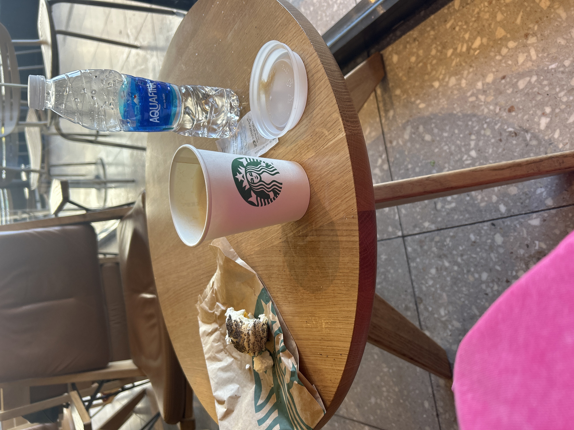 The image depicts a round wooden table at a Starbucks location in Kadıköy, Turkey. On the table are an Aquafina water bottle, a Starbucks cup with a white lid, and a pastry in a Starbucks paper bag. A portion of a chair with a brown cushion is visible on the left side of the image. A pink object, potentially part of clothing or another chair, is visible on the right side. The floor appears to be tiled in a grey pattern, and through the window in the background, more seating can be seen.
