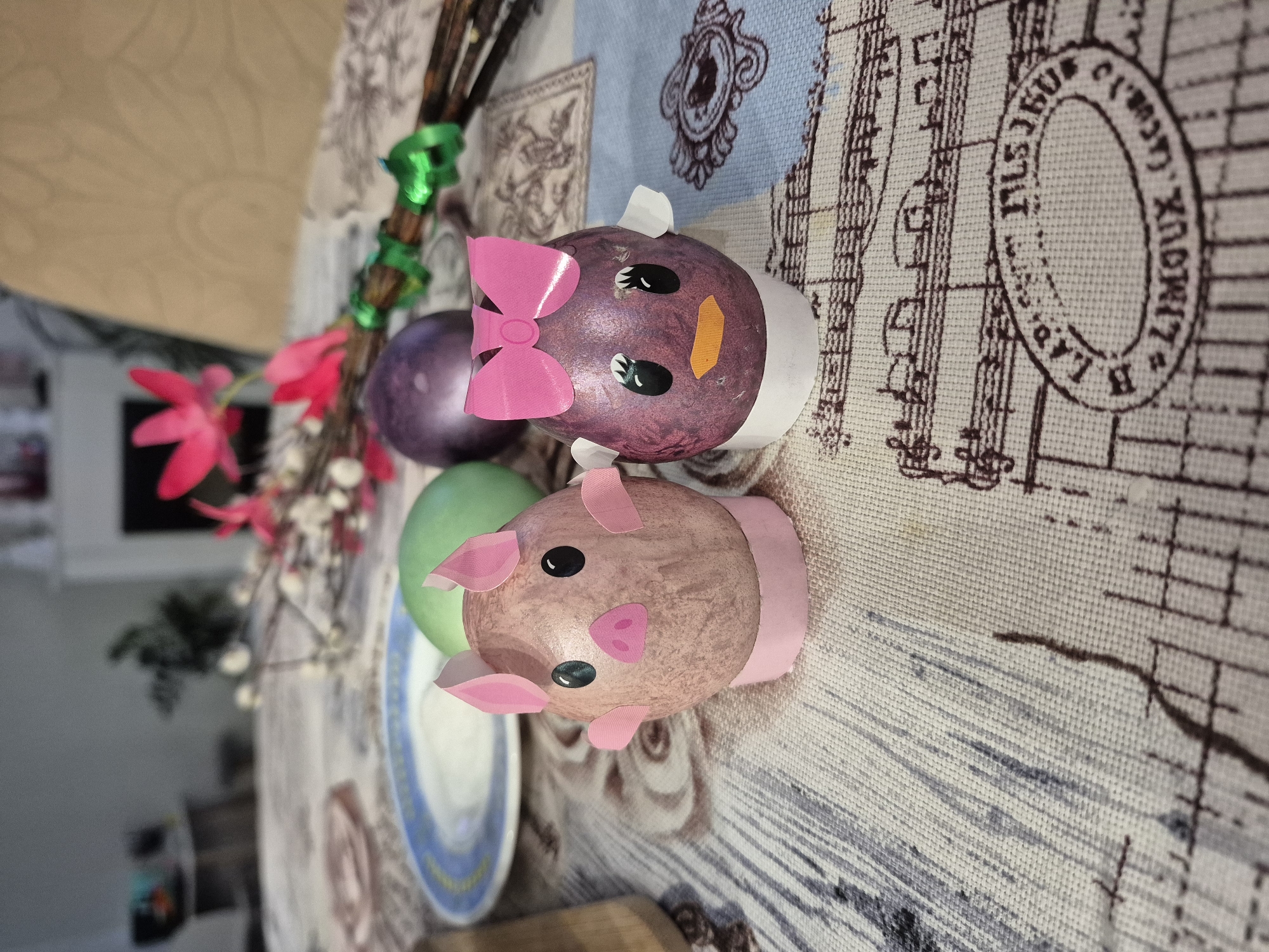 A tabletop scene features decorated eggs, including one designed as a pig and another as a bird with a pink bow, alongside dyed eggs and floral decorations. The scene is set on a tablecloth with a patterned design. The location is Chaikovsky, Russia.