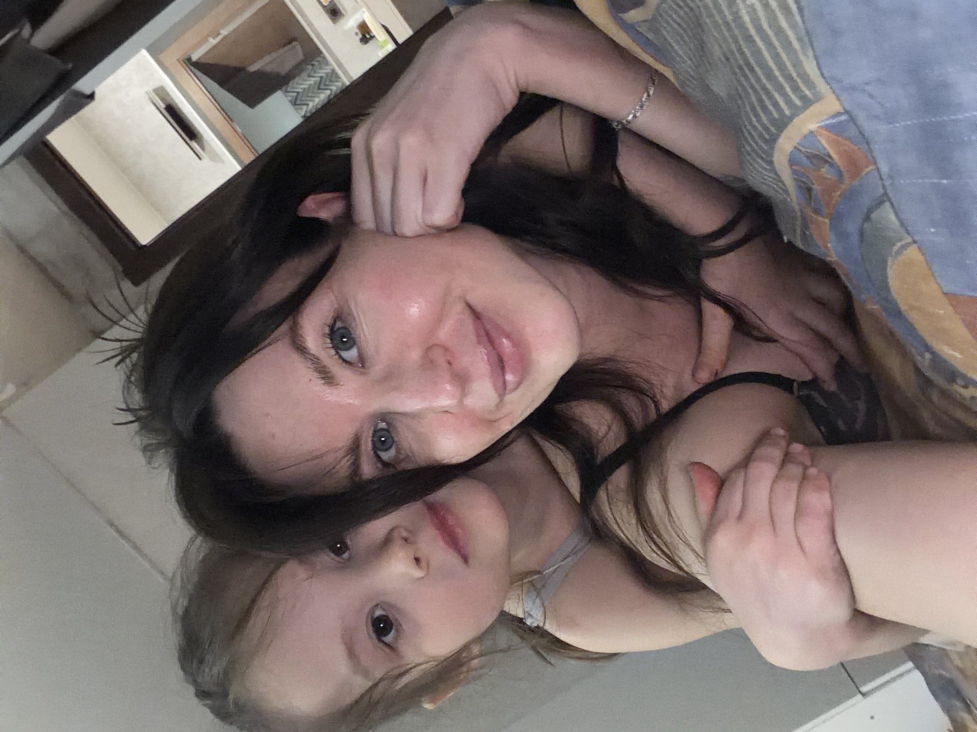 A woman and a child are lying down close to each other, possibly in a bed covered with a patterned blue and brown blanket. The woman has dark hair and blue eyes and is looking directly at the camera, smiling slightly. The child, with lighter hair, is nestled close to the woman. The location appears to be indoors. The city of Perm, Russia is not recognizable within the image.