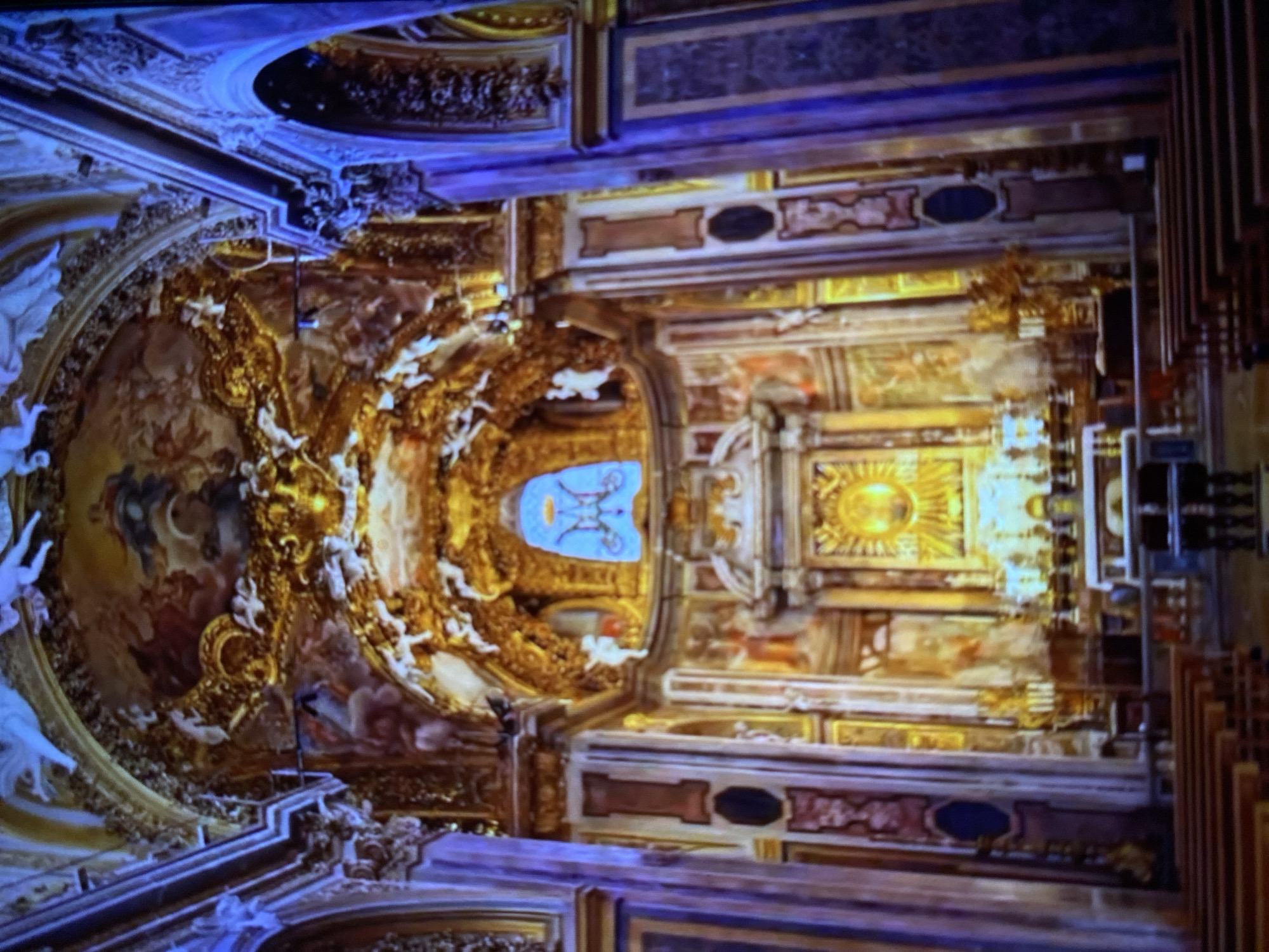 The image shows the interior of a church, likely taken from a low angle looking upwards. The ceiling is adorned with elaborate paintings, gilded ornamentation, and sculptures. There is a prominent dome section decorated with religious figures and scenes. Walls are highly ornamented with architectural elements, including detailed friezes, panels, and decorative moldings. One area of the ceiling features a golden, radiant sunburst design. A stained glass window or panel is visible in the upper center of the image. The lighting is warm and ambient, highlighting the gold details and paintings. The scene is likely located in Rome, Italy.