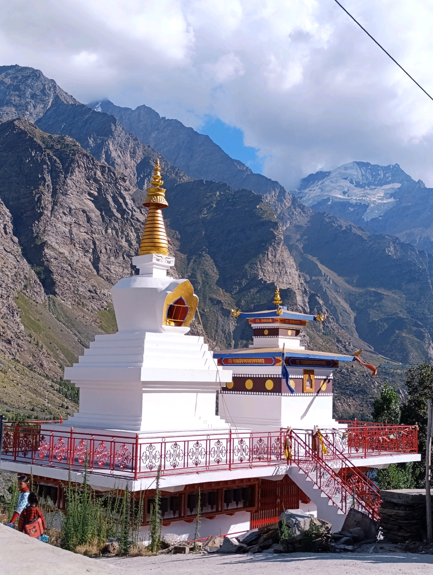 The image captures a Buddhist stupa and monastery complex situated in Gemur, India. The main stupa, a prominent white structure with a golden spire, is adorned with decorative elements and painted eyes. Adjacent to it is a smaller, colorful building with blue and white roofs, also featuring intricate designs and inscriptions. The complex is surrounded by a red and white railing, with steps leading up to an elevated platform. Below the platform, prayer wheels are visible. In the foreground, two young individuals are present, one wearing a patterned shirt and the other a red and black striped shirt with a backpack. The environment is mountainous, with rugged, rocky slopes dominating the background. Snow-capped peaks are visible in the distance, under a sky filled with white clouds. The overall scene depicts a serene religious site nestled within a dramatic natural landscape.