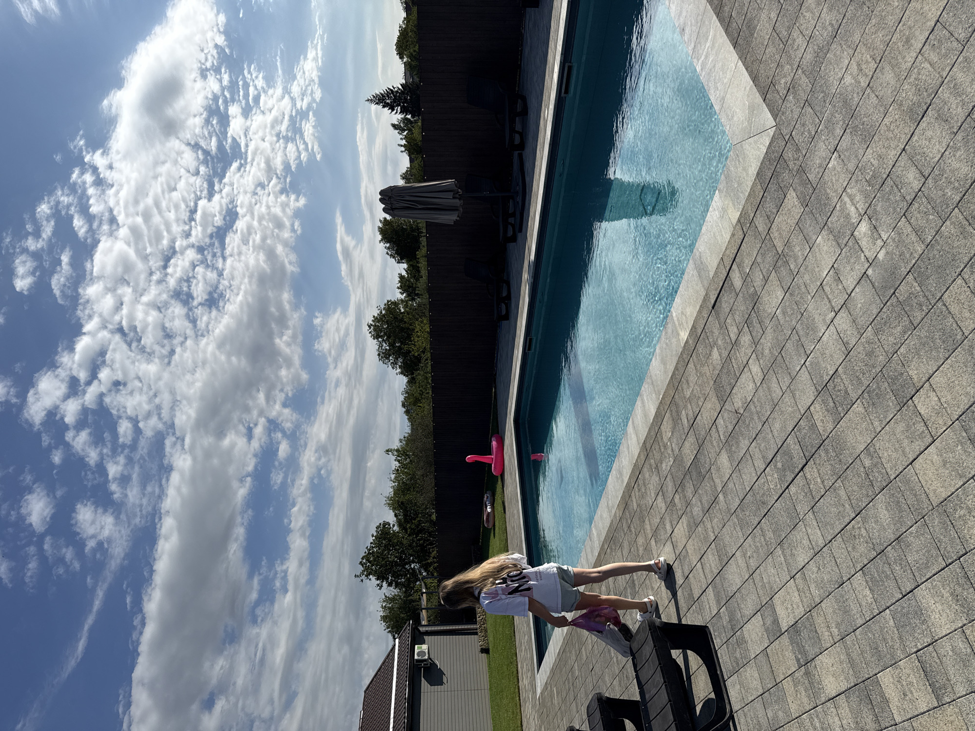 A young female with long blonde hair walks towards a swimming pool on a sunny day in Pervorechenskoye, Russia. She is wearing a white t-shirt and light-colored shorts, carrying a pink and white bag. The swimming pool is rectangular with blue water and a light-colored tiled edge. Around the pool are several dark-colored lounge chairs and a folded umbrella. The surrounding area is paved with grey interlocking stones. In the background, there is a dark wooden fence, trees, and a small building with a brown roof. The sky is blue with scattered white clouds.