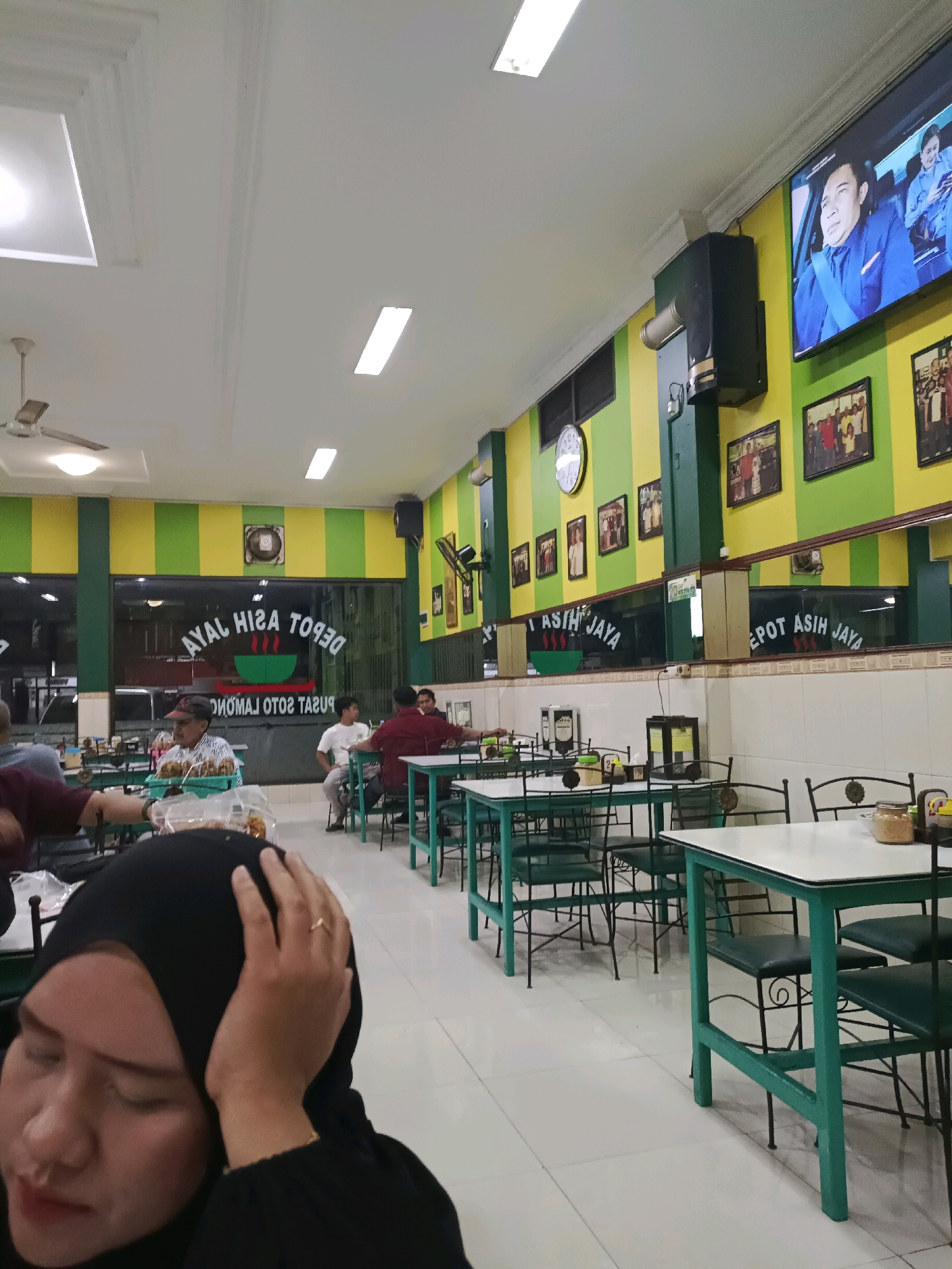 The image shows the interior of a restaurant, possibly "Depot Asih Jaya" based on the signage visible through the window. The restaurant has a distinctive yellow and green striped decor on the walls. Several tables and chairs are arranged in the dining area, with some occupied by customers. In the foreground, a woman wearing a black hijab is visible, with her hand on her head. In the background, a television screen displays a scene from a program. The restaurant appears to be located in a city in Indonesia, possibly in Dapur based on the textual hints. The overall impression is of a casual dining establishment.