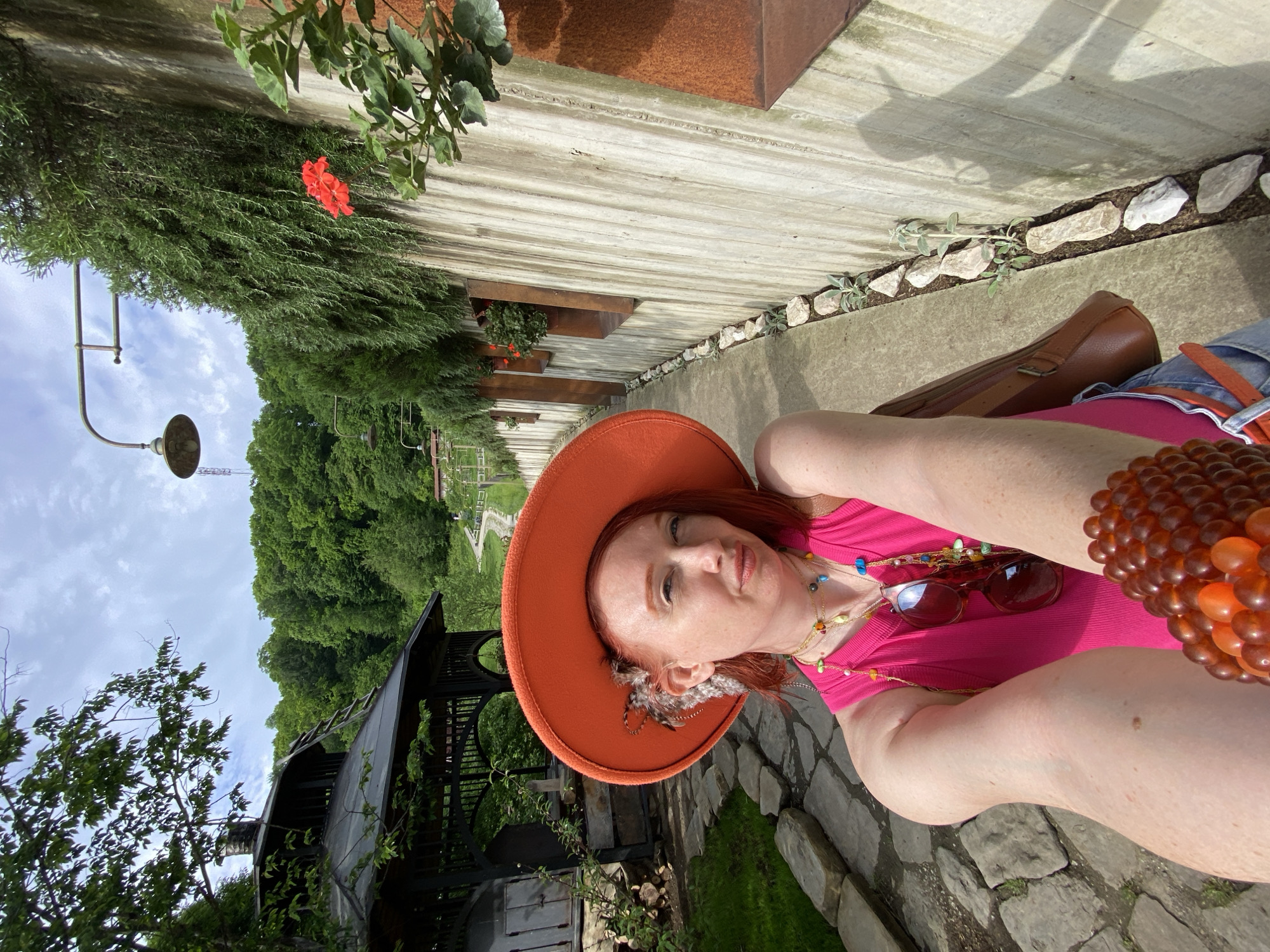 A person with red hair and an orange hat is in an outdoor setting. She is wearing a pink top, jewelry, and sunglasses. Beside her is a brown bag. Behind her is a building with greenery, a concrete wall with plants in it, a walkway with stones, and a gazebo. In the background, there is a tree-covered landscape and a cloudy sky. I have no information to suggest this image was taken in Прогресс, Russia.