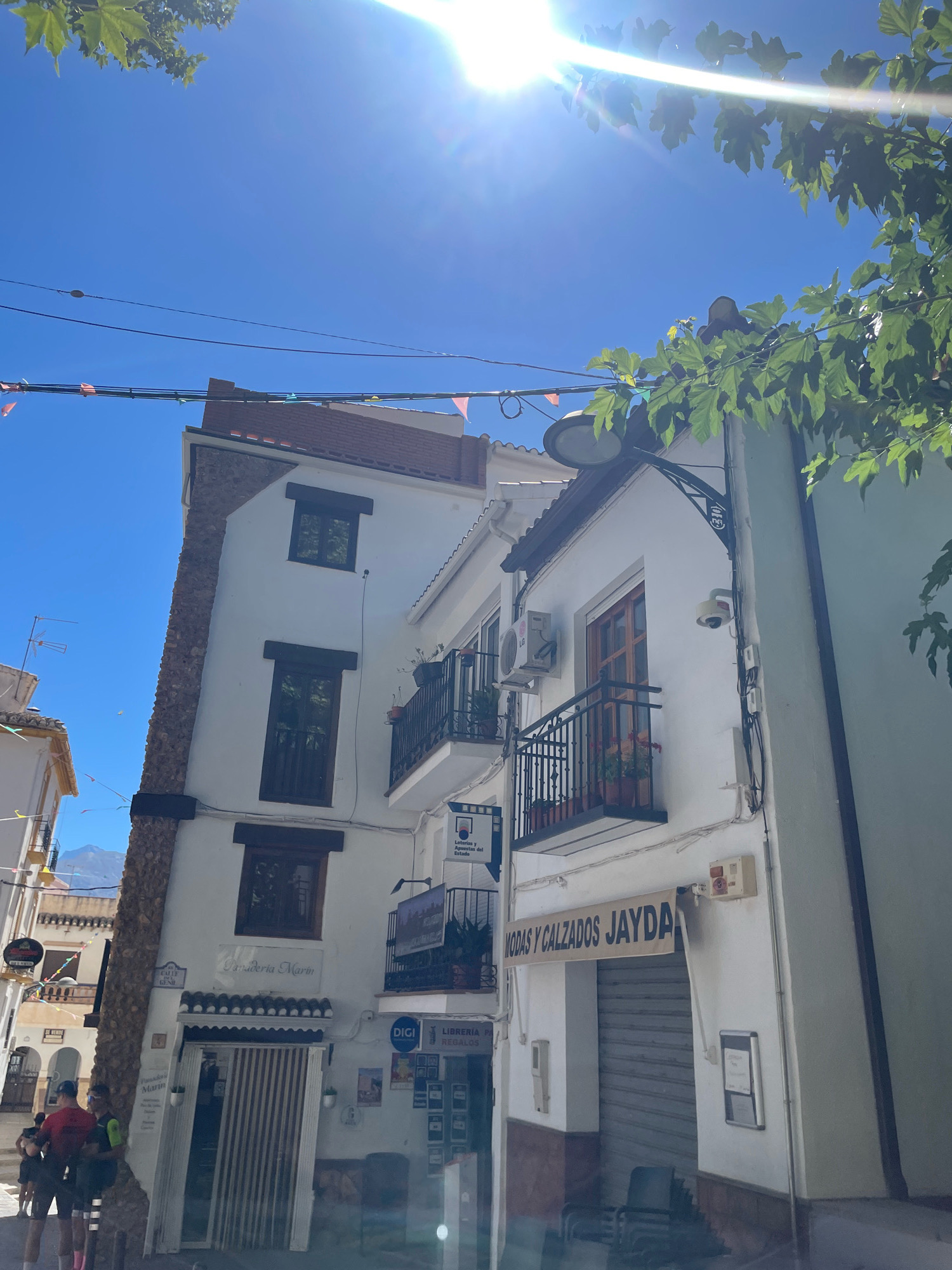 The image shows a street scene in Güéjar Sierra, Spain. In the foreground, a white building with a brown trim is visible, featuring a storefront with a closed metal shutter and an awning that reads "MODAS Y CALZADOS JAYDA". Above this, a balcony with black railings and potted plants is present, along with an air conditioning unit and a security camera. To the left, another building is partially visible with a stone-clad section and several windows. A street sign reading "CALLE GENIL" is mounted on this building. In the lower left of the image, a group of four people, dressed in athletic wear, are walking. The sky is clear and blue with the sun prominently shining, casting a lens flare and highlighting green foliage from a tree in the upper right corner. Decorative banners are strung across the street. In the background, the silhouette of mountains can be seen.