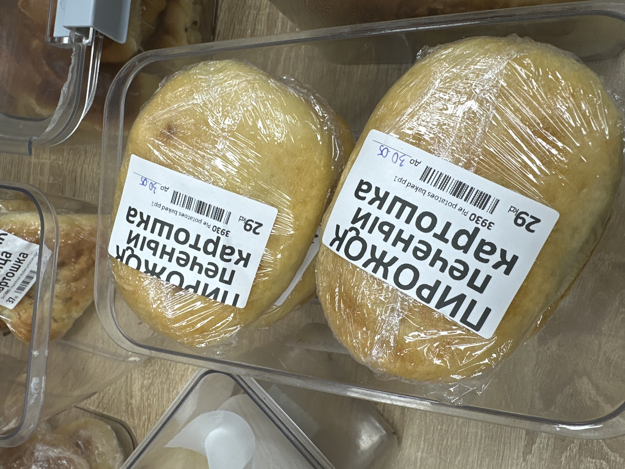 Here is a forensic description of the image:
The image presents a close-up, overhead view of a plastic container holding two baked potato pies wrapped in clear plastic film. Each pie has a white label affixed to it, printed in Cyrillic text, presumably Russian, along with a price of "29kd" and a date "до 30.05". Text reads: "ПИРОЖОК печеный картошка". A barcode is also present on the label. The text below the price and date, states "3930 Pie potatoes baked pp1"
Part of another container with baked goods, also wrapped in plastic, is visible to the left of the main container. The surface underneath the containers appears to be a light-colored wood grain.
There is nothing present in the image that suggests the location in the city of Bac Nha Trang Ward, Vietnam.