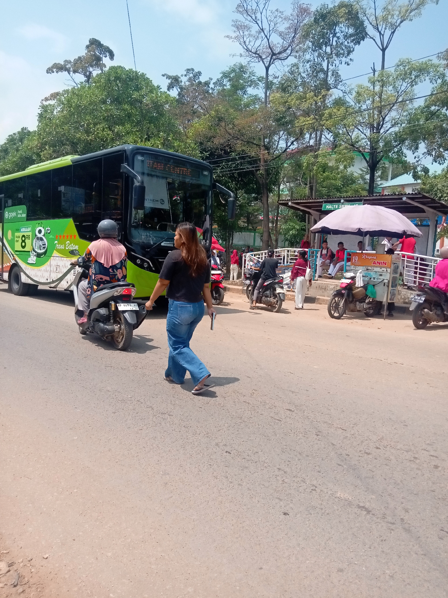 A person walks across a paved road in front of a green and white bus and a bus stop. The bus stop has a white canopy and several people are sitting under it. There are motorcycles parked near the bus stop. The scene appears to be in Batam, Indonesia, as indicated by the text "Trans Batam" on the bus.