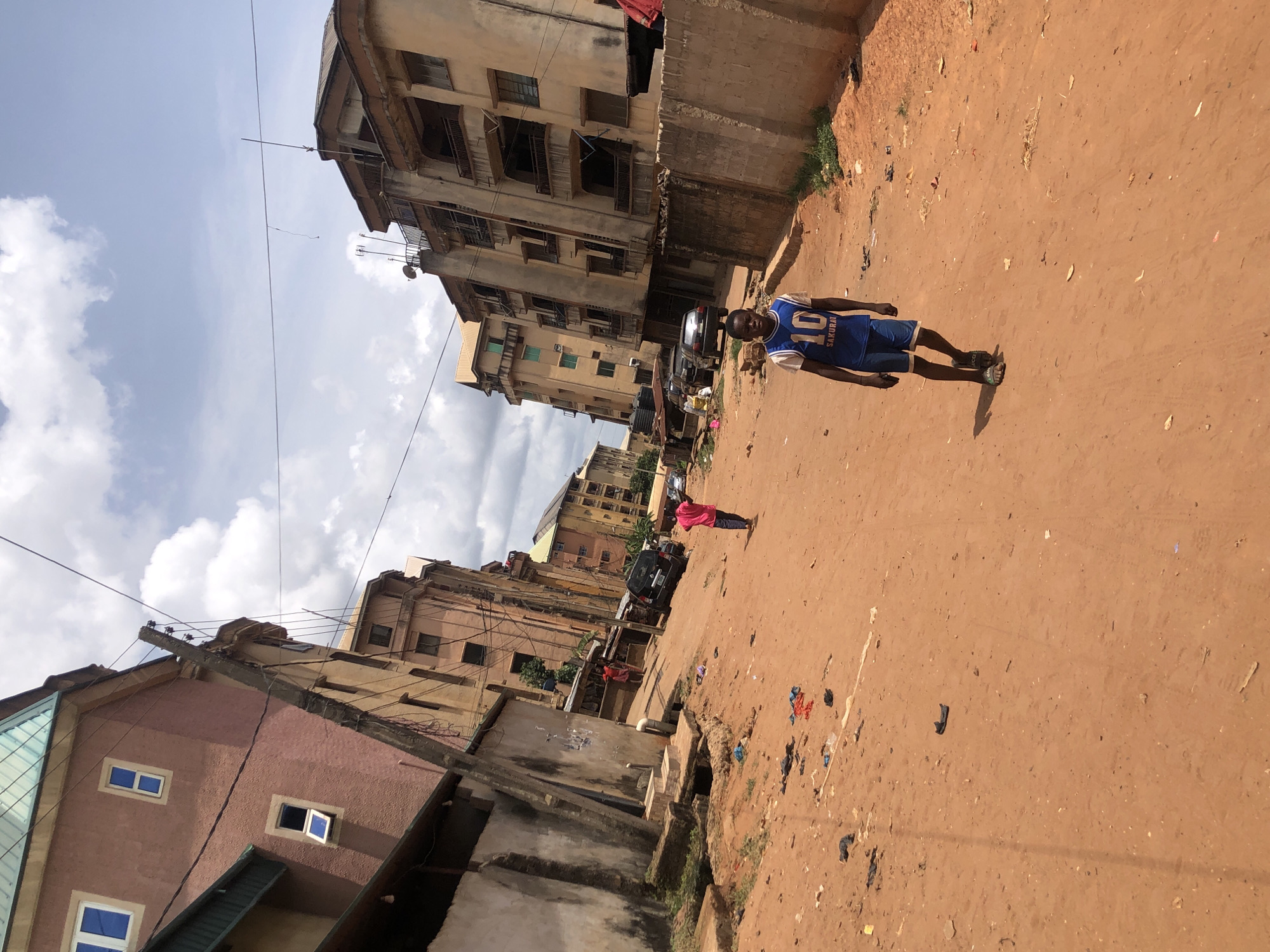The image depicts a street scene in Onitsha, Nigeria. The road is unpaved, consisting of reddish-brown soil with scattered debris. A young man wearing a blue jersey with the number 10 and the name "Sakurai" on the back stands on the right side of the road, facing forward. In the background, there are several multi-story residential buildings with faded paint and balconies. A woman wearing a red shirt is standing farther down the street near parked vehicles. Power lines are visible overhead against a cloudy sky. The scene appears to be a typical residential area with signs of urban decay.