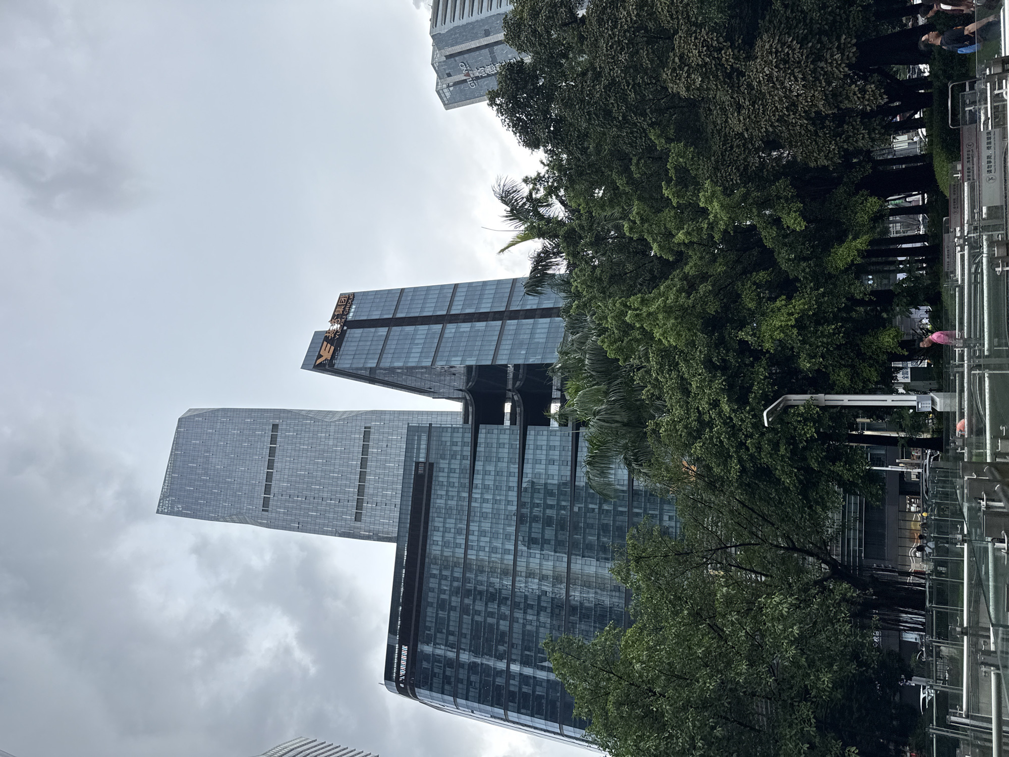The image shows a cityscape in Tianhe District, China, with modern skyscrapers and lush trees. The sky is overcast, suggesting it might be a cloudy or rainy day. The buildings are tall and feature glass facades, reflecting the grey sky. One building has visible text in Chinese characters and "YUEXIU GROUP" written in English. Another building has "GRAND" written on it. Several people are visible in the background, near what appears to be an entrance or walkway with metal railings and signs. The scene is vertical, with the top of the frame showing the sky and the bottom showing parts of the buildings and trees. The overall impression is one of an urban environment with significant green spaces integrated into the architecture.