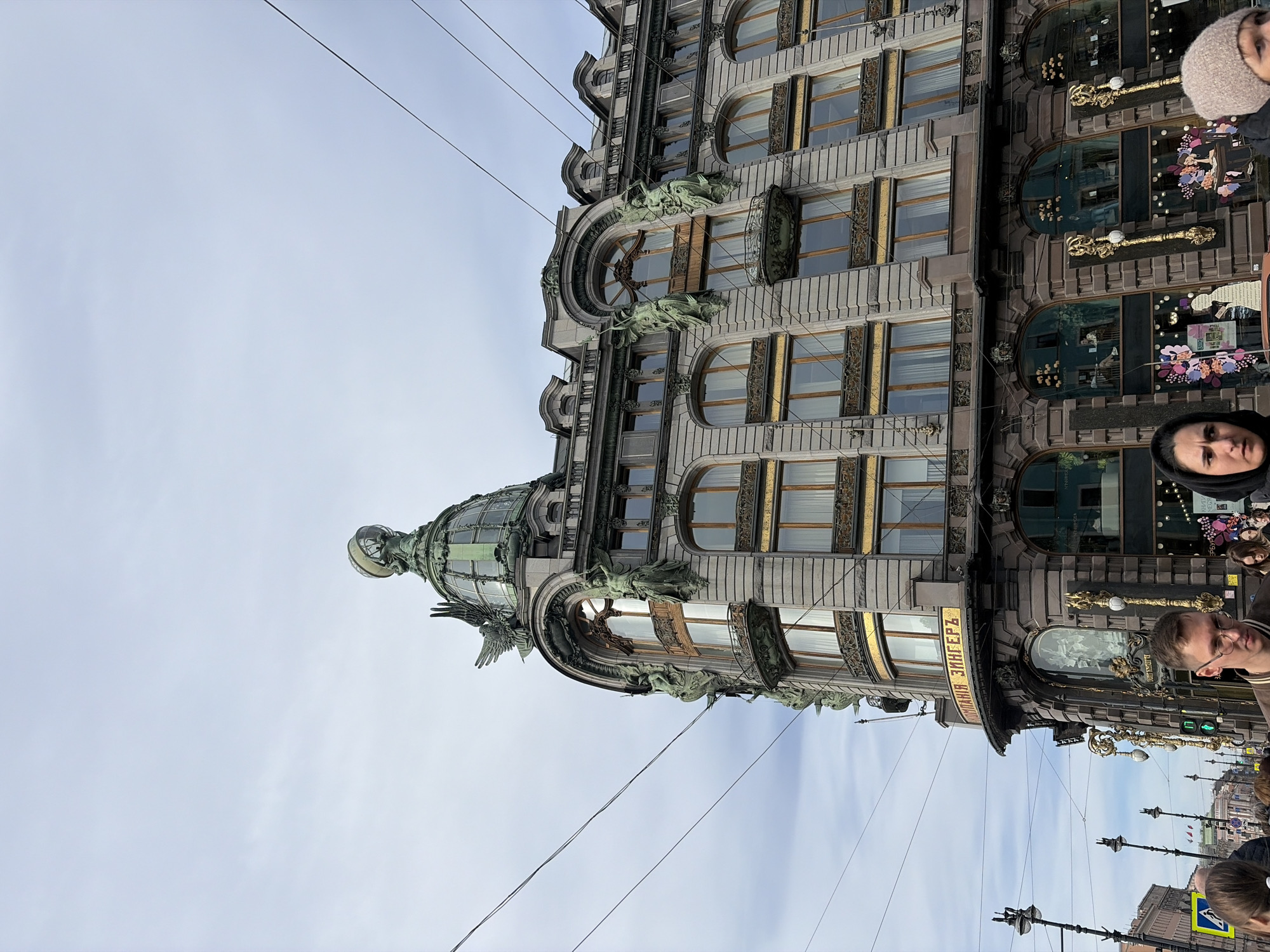 The Singer House, an ornate architectural landmark in Saint Petersburg, Russia, dominates the scene. The building's facade, featuring multiple arched windows with intricate gold trim and decorative bronze sculptures, is partially visible against a cloudy sky. Power lines stretch across the sky in front of the building. A crowd of people is visible near the base of the building, including individuals with faces and upper bodies partially visible. Street lights and additional buildings are visible in the background, providing context for the urban setting.