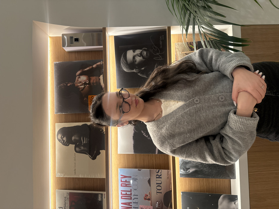 A woman with glasses and a gray cardigan stands in front of a record shelf. Several record albums are displayed, including "Silence," "Sade Love Deluxe," "Lana Del Rey Honeymoon," "Travis Scott," and "Scorpion." The shelves are illuminated with warm light, creating a cozy ambiance. A plant's fronds are partially visible in the upper right corner, adding a touch of nature to the scene. The woman's hands are clasped.