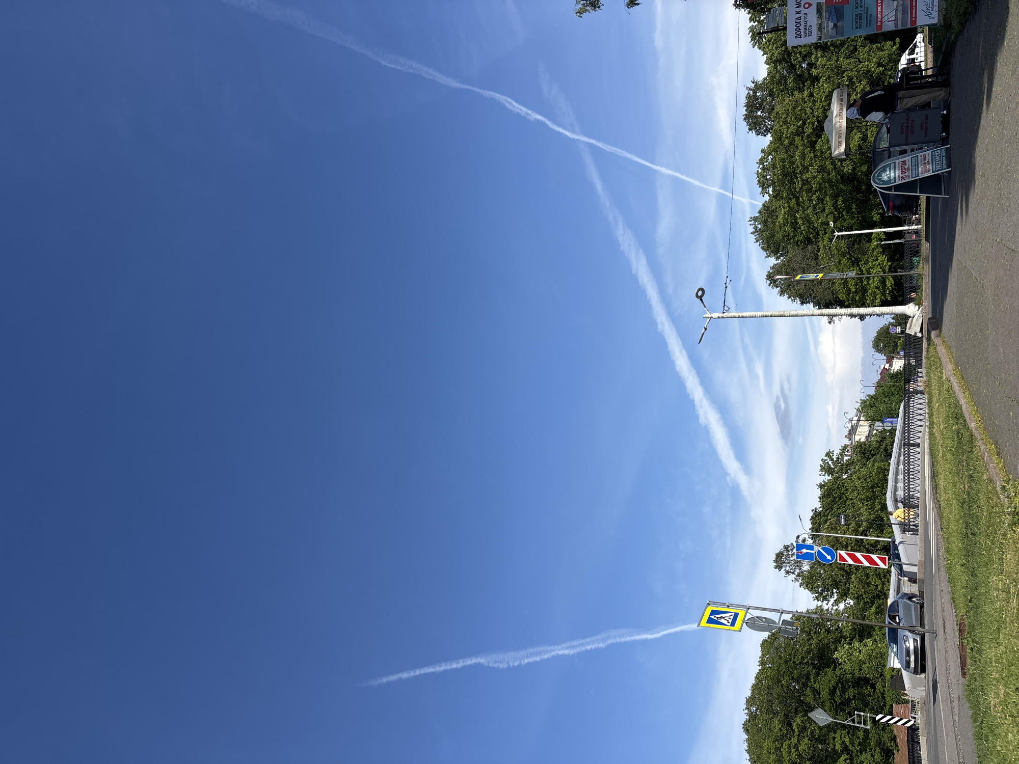The image depicts a bright, sunny day in Kronstadt, Russia, with a clear blue sky traversed by several airplane contrails. In the foreground, a paved walkway borders a grassy area. A street with cars and traffic signs runs through the scene, with a pedestrian crossing visible. Lush green trees line the background, interspersed with buildings. A small kiosk or stand is set up on the walkway, with a person standing nearby. Several advertising signs are present, some in Russian. The overall impression is a typical urban scene on a pleasant day.