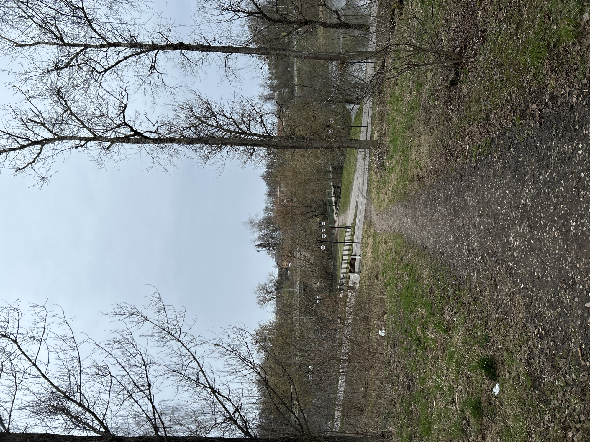 The image shows a gravel path in Klin, Russia, likely along the Sestra River. The path is flanked by grass and bare trees. There are lamp posts and benches visible. The sky is overcast. No people are visible. There is some litter on the ground.