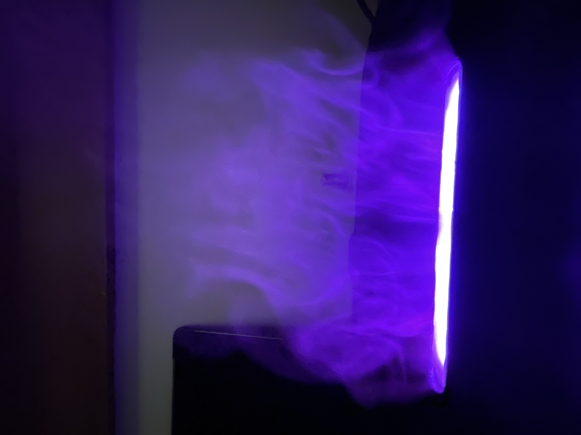 The image displays a close-up, low-angle shot of a dark, flat surface with a bright, horizontal light source emitting purple illumination. Wisps of white mist are visible, swirling upwards from the light source and catching the purple glow. The background is a gradient of dark to light gray. The scene appears to be indoors, with a dark object to the left and a dark ceiling or shelf above. There is no indication of the city of Da Nang, Vietnam, or any specific urban environment.