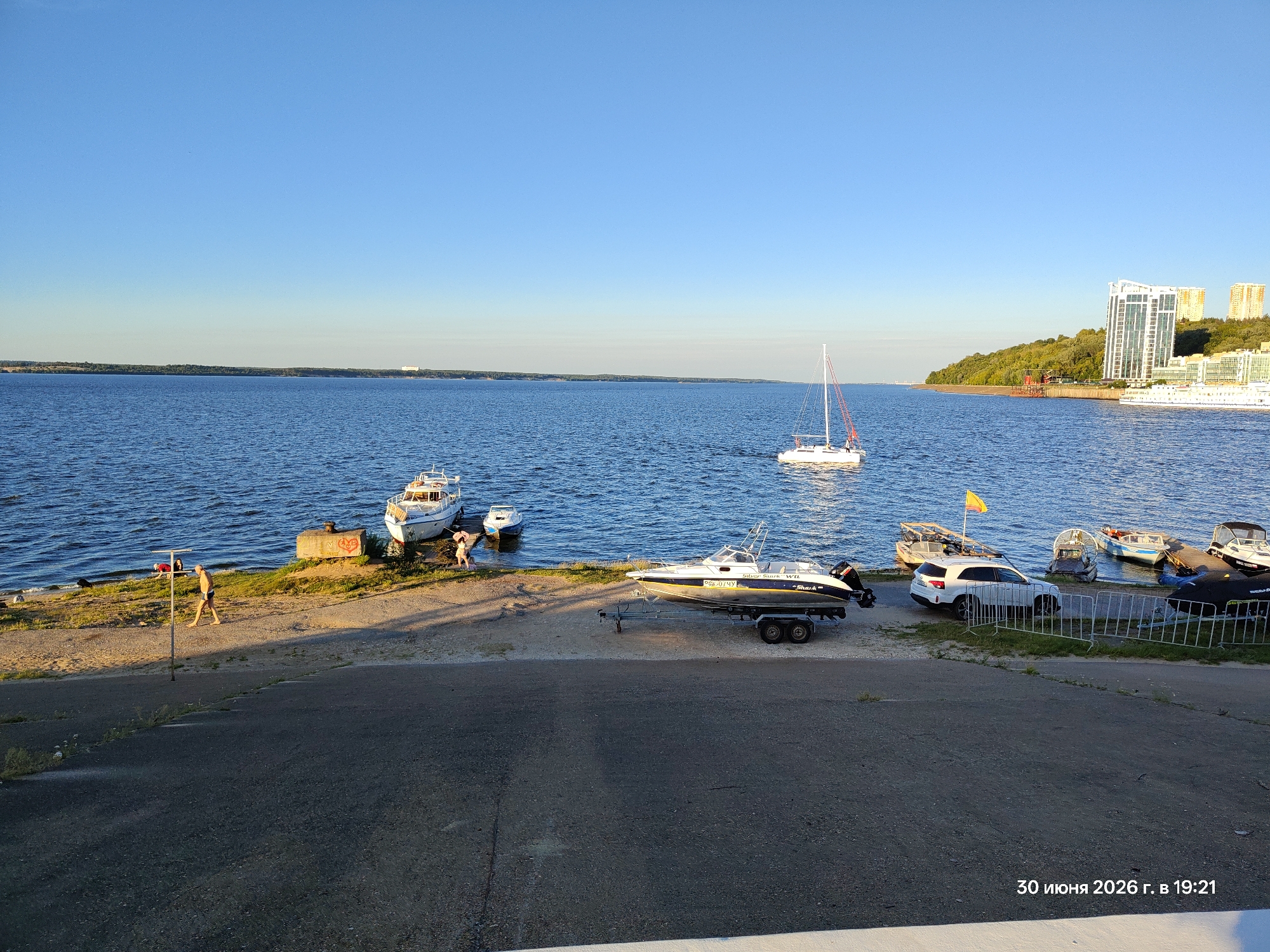 A serene waterfront scene unfolds under a clear blue sky. The deep blue water of a wide river or lake stretches towards a tree-lined horizon, dotted with several boats. In the foreground, a paved ramp slopes down towards the water's edge, where a few individuals are visible. One person, a man in dark shorts, walks along the grassy bank, bathed in the warm light of a late afternoon or early evening sun. To his right, a white motorboat rests on a trailer, its sleek design gleaming. Further along the bank, several other boats are moored, some appearing more rustic and others more modern.
In the middle ground, a white sailboat with a red-tipped mast glides across the water. Beyond it, on the far shore, a dense green forest meets the water, with modern high-rise buildings peeking out from behind the trees, suggesting a city or urban area is nearby. The sun casts long shadows, indicating it is likely late afternoon or early evening. The overall atmosphere is peaceful and tranquil, evoking a sense of leisure and a pleasant summer day.
The lower right corner of the image contains visible text in Russian: "30 июня 2026 г. в 19:21", which translates to "June 30, 2026 at 19:21". This indicates the date and time the photograph was taken.