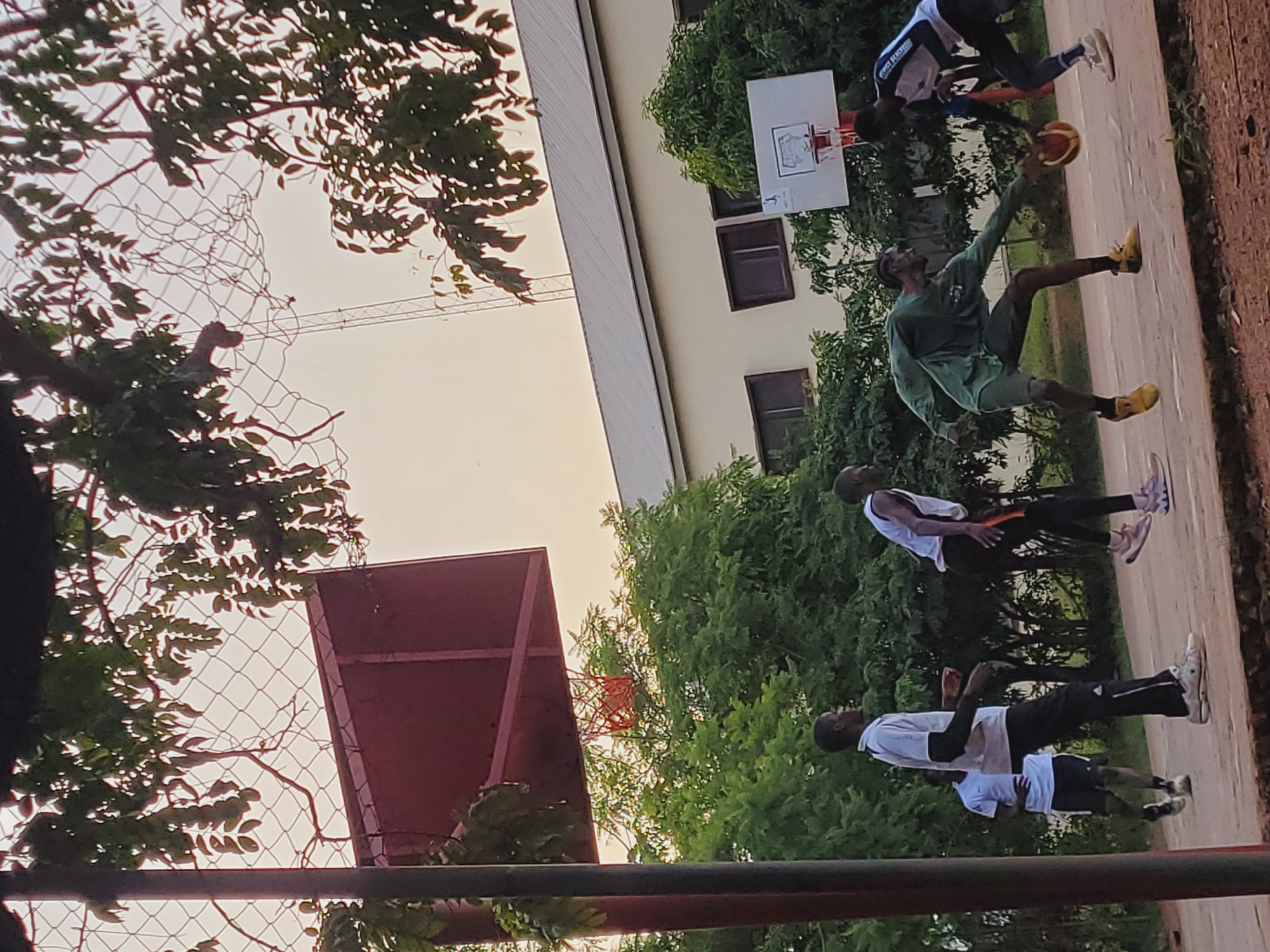 In Bukuru, Nigeria, on an outdoor basketball court with a chain-link fence and trees, several young men are playing basketball. A basketball is in play between two players in the foreground. One player in a green long-sleeved shirt and grey shorts reaches for the ball, while another player in a white and blue jersey and black pants defends. In the background, two other players in white shirts and black pants are also present on the court. A building with a metal roof and windows is visible behind the court, and the sky is overcast.