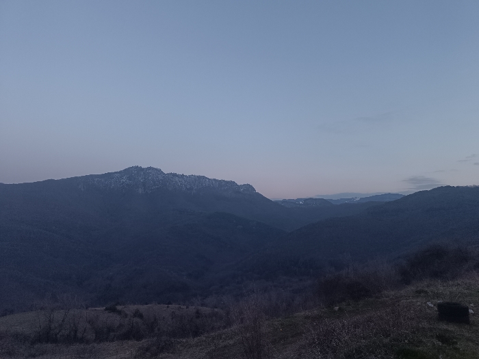 A panoramic landscape photograph depicts a mountainous region under a twilight sky, specifically located in Oreshets, Bulgaria. The sky occupies the upper two-thirds of the frame, transitioning from a light blue-gray at the zenith to a faint pinkish-orange hue at the horizon. A prominent mountain range stretches across the mid-ground, its peaks, particularly on the left and center, exhibiting patches of snow or frost. The mountains are heavily vegetated with dark, indistinct trees, appearing in deep blue-gray tones due to the low light. Further mountain ranges recede into the distance on the right. The foreground consists of an uneven, sparsely vegetated slope covered in dry, brown-gray ground and scattered bare deciduous shrubs or small trees. A dark, undefined object is present in the lower right corner of the foreground. No human figures, animals, or discernible structures are visible. The lighting conditions are consistent with dusk or dawn, producing a subdued palette and long shadows across the terrain.