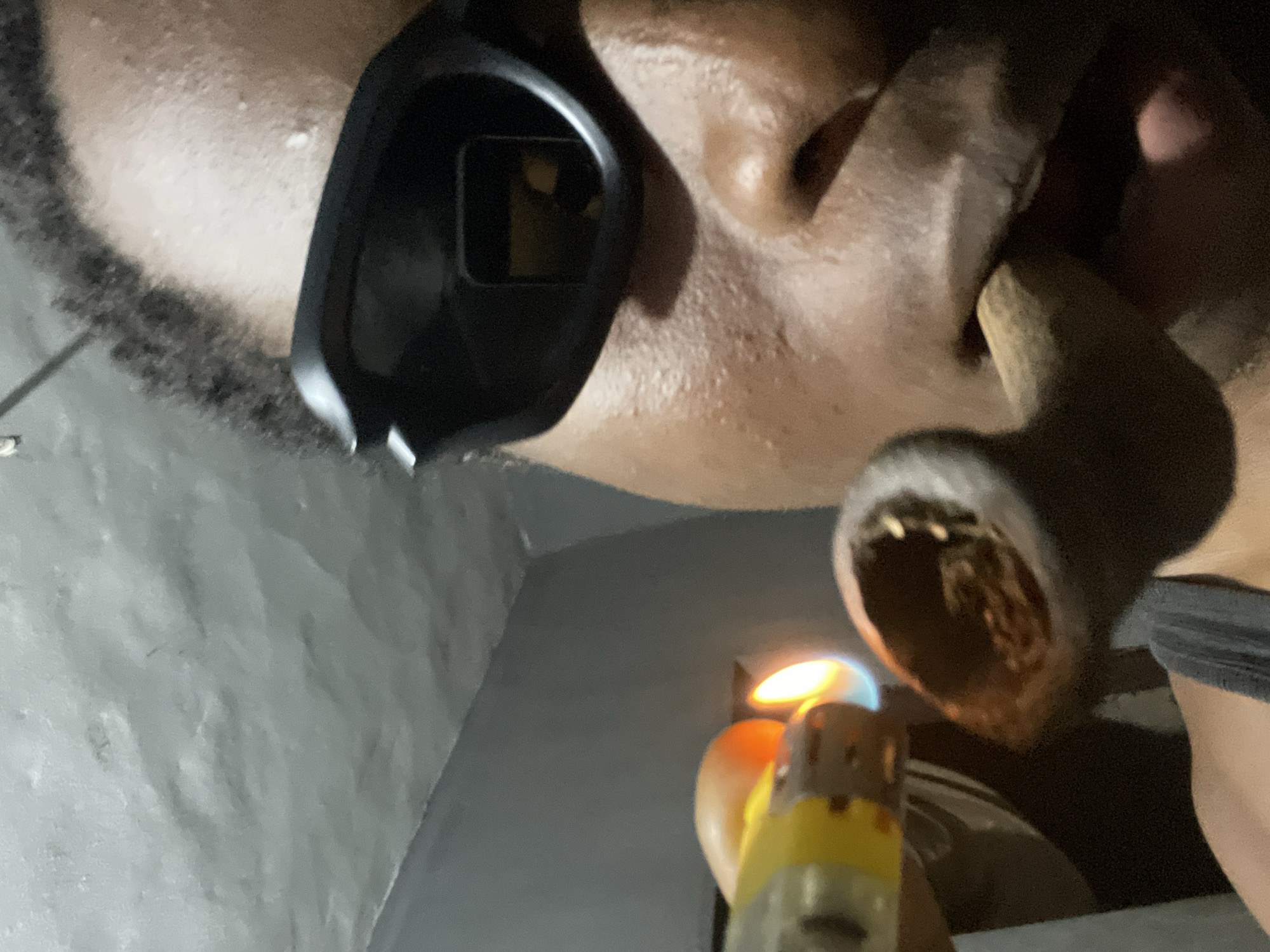 The image, set in Obosi, Nigeria, presents a close-up, low-angle perspective of an individual lighting a pipe. The person, whose skin appears dark, has short, dark hair or stubble visible on the side of their head. They are wearing a dark, single-lens eyepiece or eyewear on the left side of their face. A pipe, crafted from a dark, natural material such as wood, is held near the face, with its bowl containing a packed, dark substance. A yellow and silver lighter is actively producing an orange and blue flame, which is being applied directly to the contents of the pipe bowl, indicating the act of smoking. The background is a grey, textured surface, likely a wall, suggesting an indoor or enclosed environment with dim lighting. A portion of a dark grey garment is also visible on the right. The lighting conditions imply it is either nighttime or a poorly lit interior. No emotions are discernible, and no text is visible within the frame.