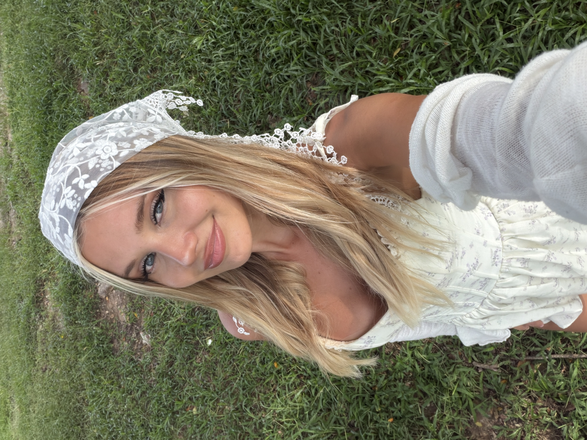 In a grassy outdoor area, a young woman with blonde hair and blue eyes is taking a selfie. She is wearing a white floral dress and a white lace headscarf. Her right arm is extended towards the camera, and she is wearing a white arm warmer. The background is composed of green grass, with some patches of dirt visible. The lighting suggests it is daytime.