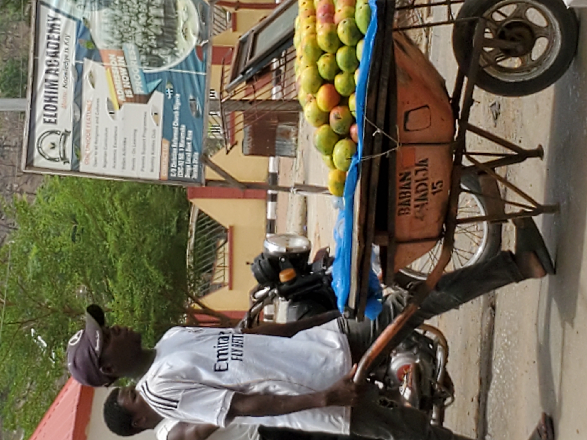 The image shows a man driving a three-wheeled vehicle loaded with mangoes. Text on the vehicle reads "BABAN HADIJA 15". Behind the vehicle is a sign for Elohim Academy that states “Admission is now in Progress”, and the school's motto "Knowledge is Key". Two men are visible, one driving and one standing beside the vehicle. The scene appears to be in an urban area with a variety of buildings and trees visible. The location is the city of Mararaba, Nigeria.