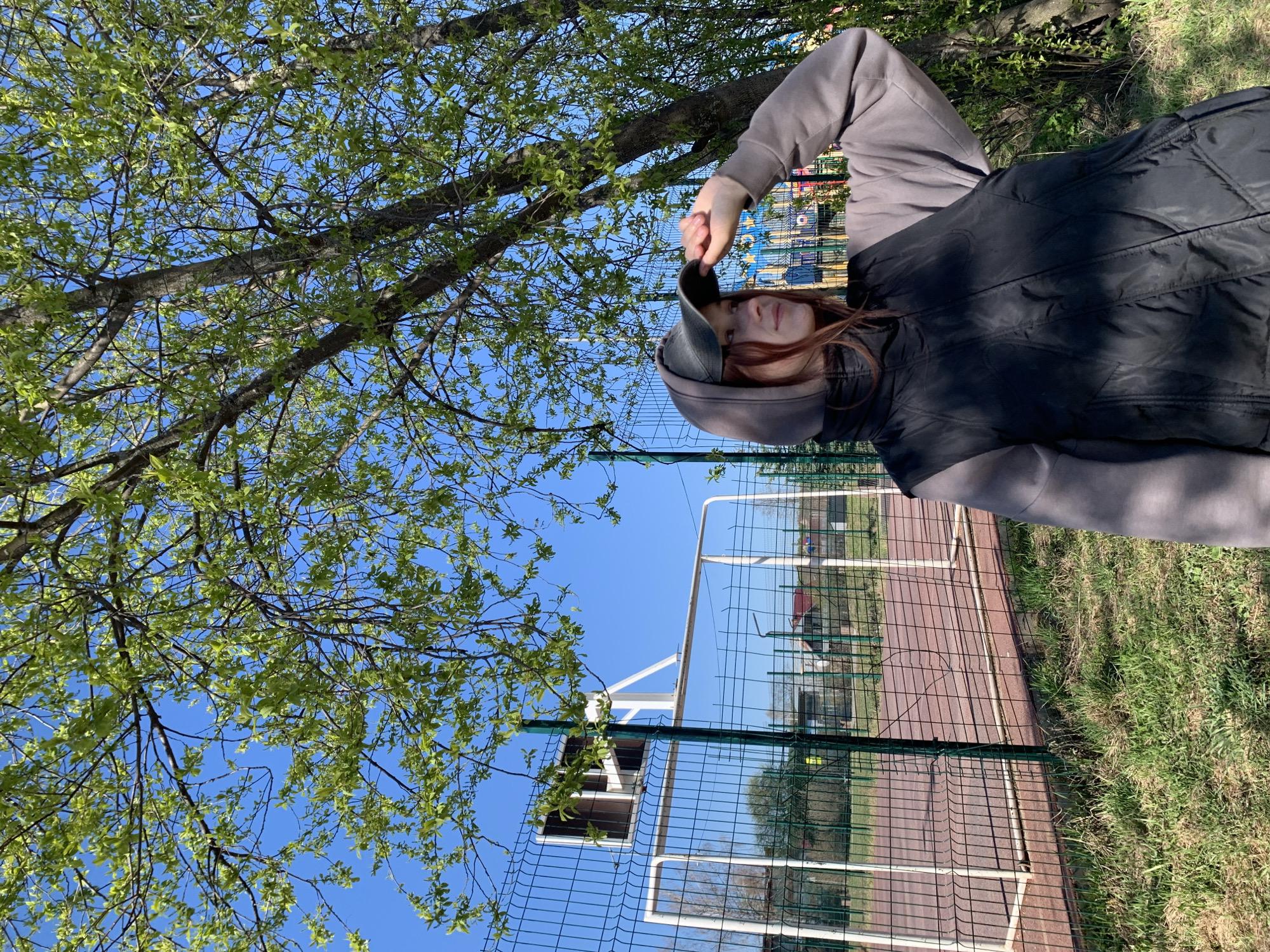 A photograph taken in Фоки, Russia, presents a young person standing outdoors next to a chain-link fence and a tree. The individual, likely a young woman, is wearing a gray hooded sweatshirt and a black vest. She is holding a gray hat above her head. The tree to her left has light green leaves, indicating it is likely springtime. In the background, visible through the fence, there's a red and tan track, possibly a running track or other sports area. Beyond the track, there appear to be buildings or structures. The sky is clear and blue. The photograph has been rotated counterclockwise.